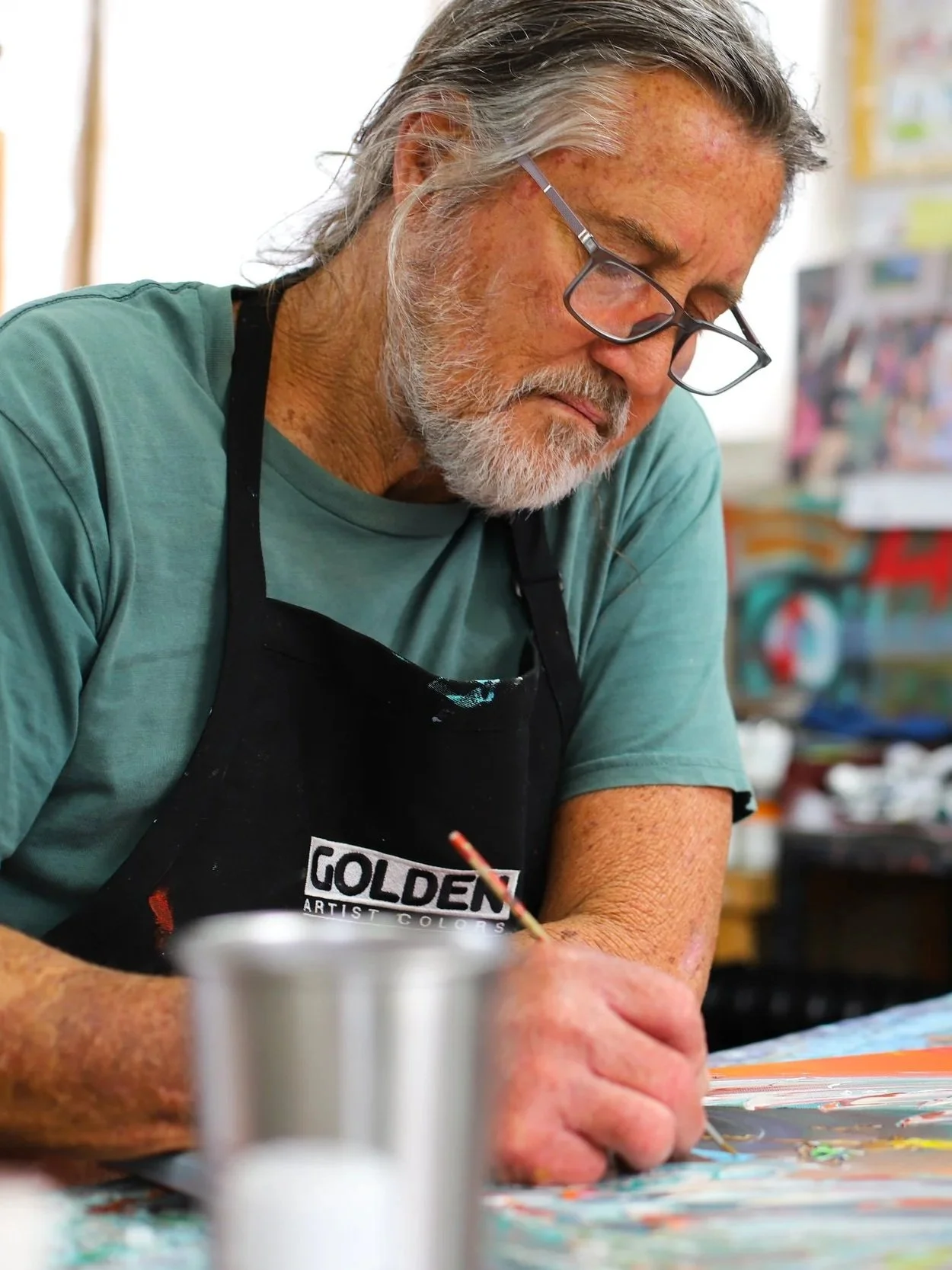 An senior man with gray hair and beard, wearing glasses and a black apron, painting with a small brush in an art studio.