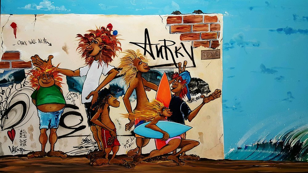 Colorful graffiti mural of cartoon-style characters with wild hair, some holding surfboards, on a wall with graffiti and a blue sky background.