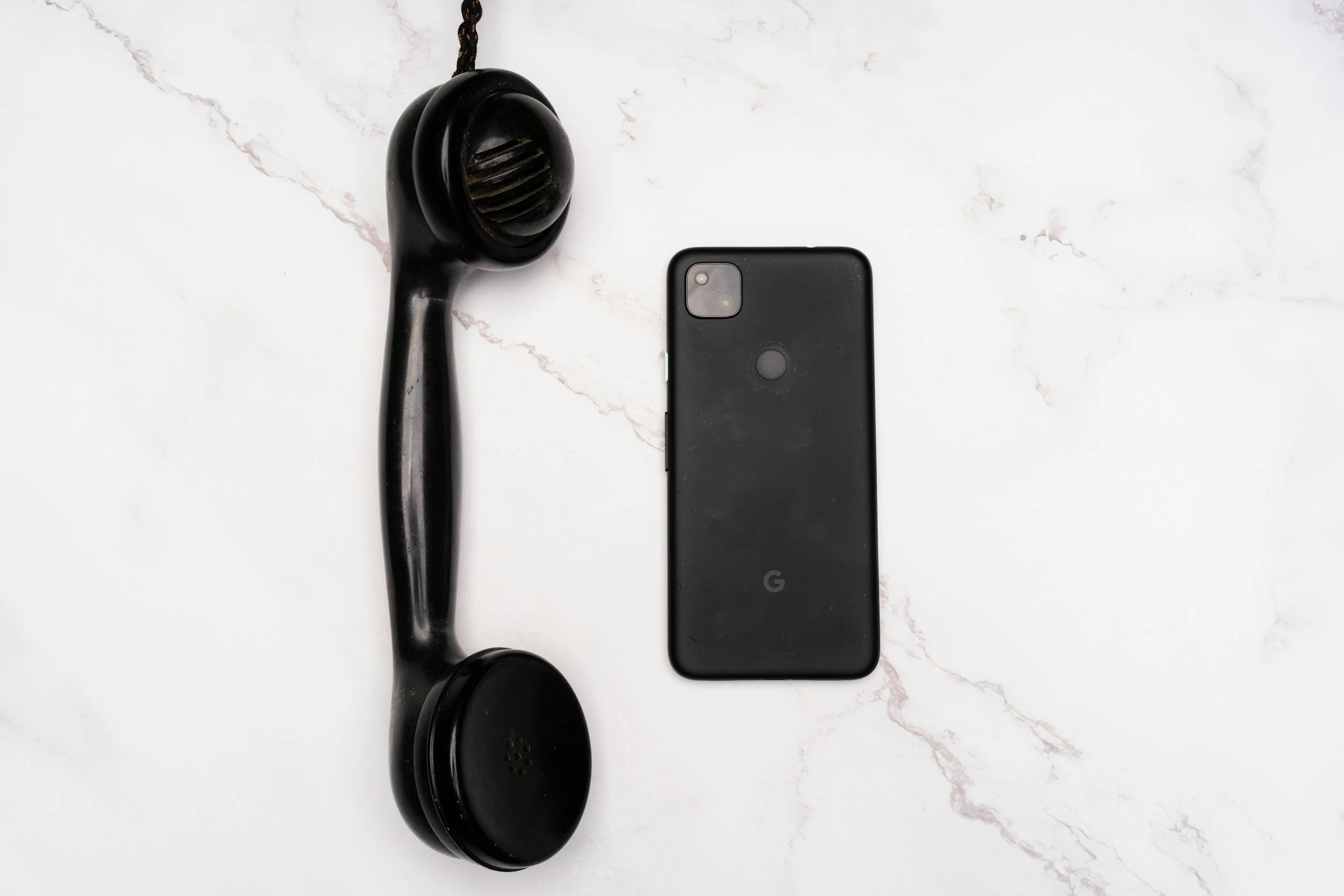 A black analog telephone receiver and a black Google Pixel smartphone on a white marble surface.