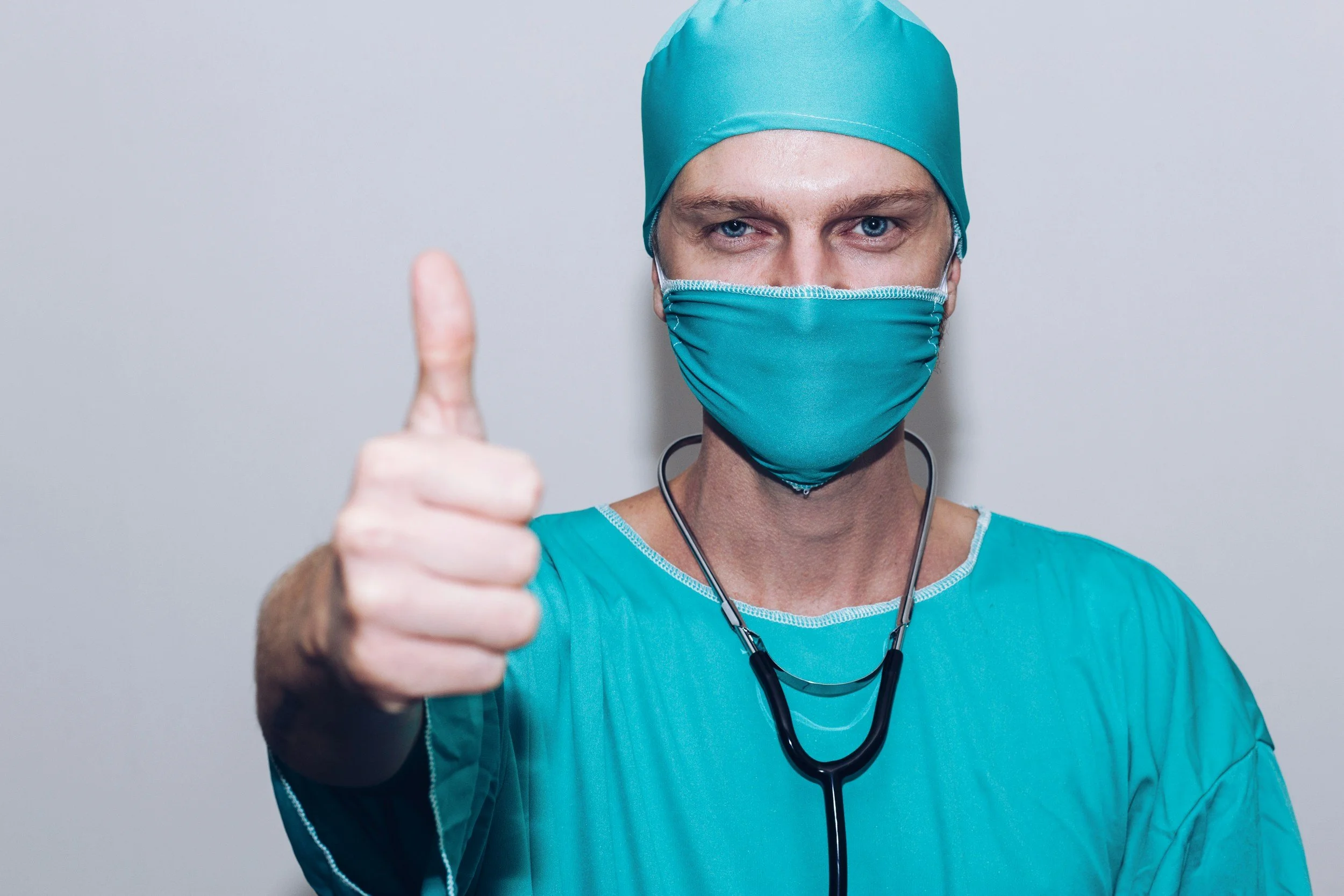 A male healthcare professional wearing teal scrubs, a surgical cap, and a face mask, giving a thumbs-up gesture.