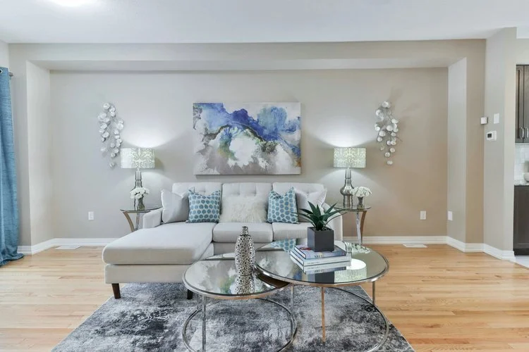 Modern living room with white sofa, abstract wall art, two table lamps, decorative pillows, glass coffee tables, a rug, and hardwood floors.