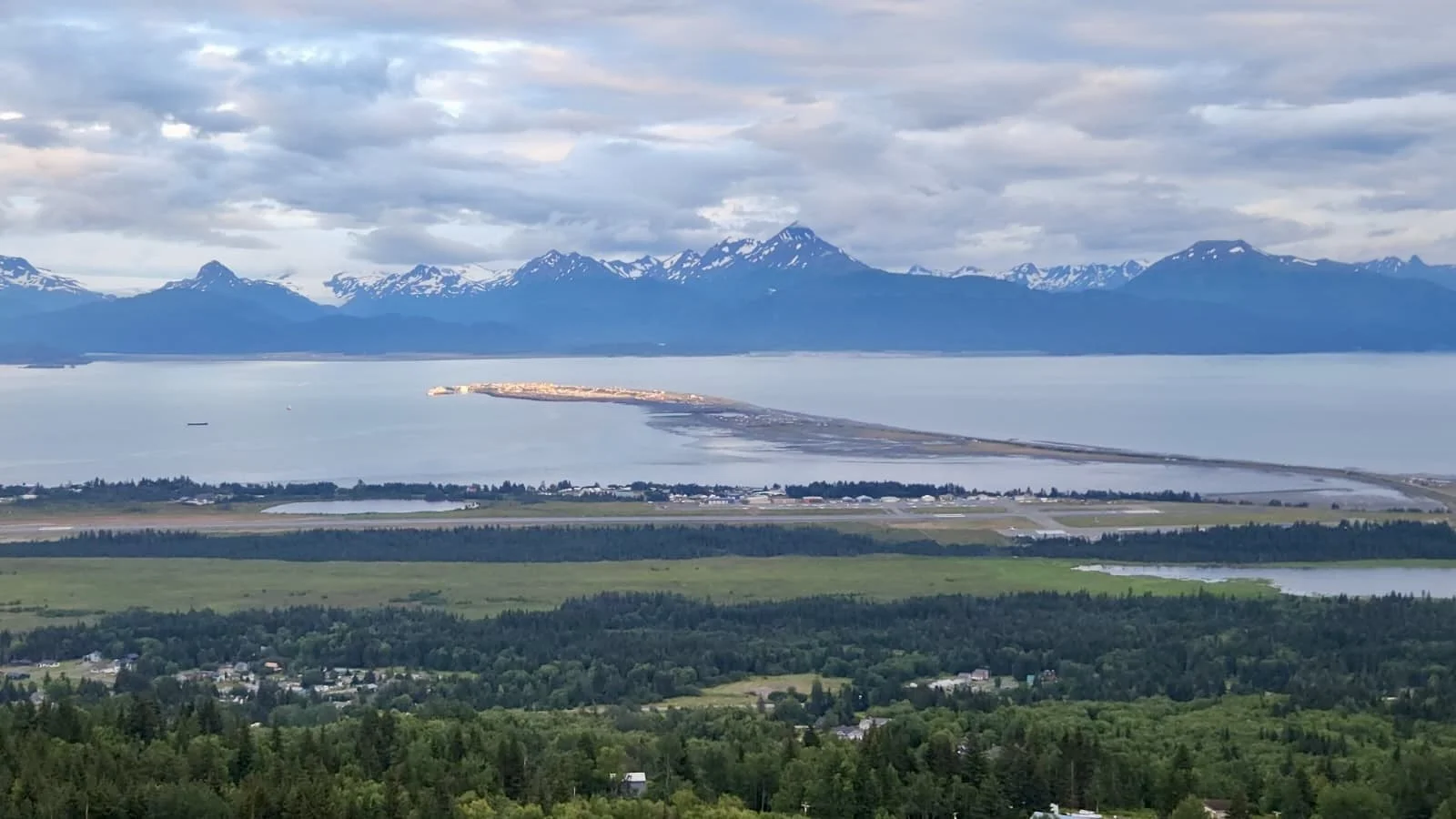 Scenic view of a large body of water with mountains in the background, a small island or peninsula in the water, and green fields and trees in the foreground.