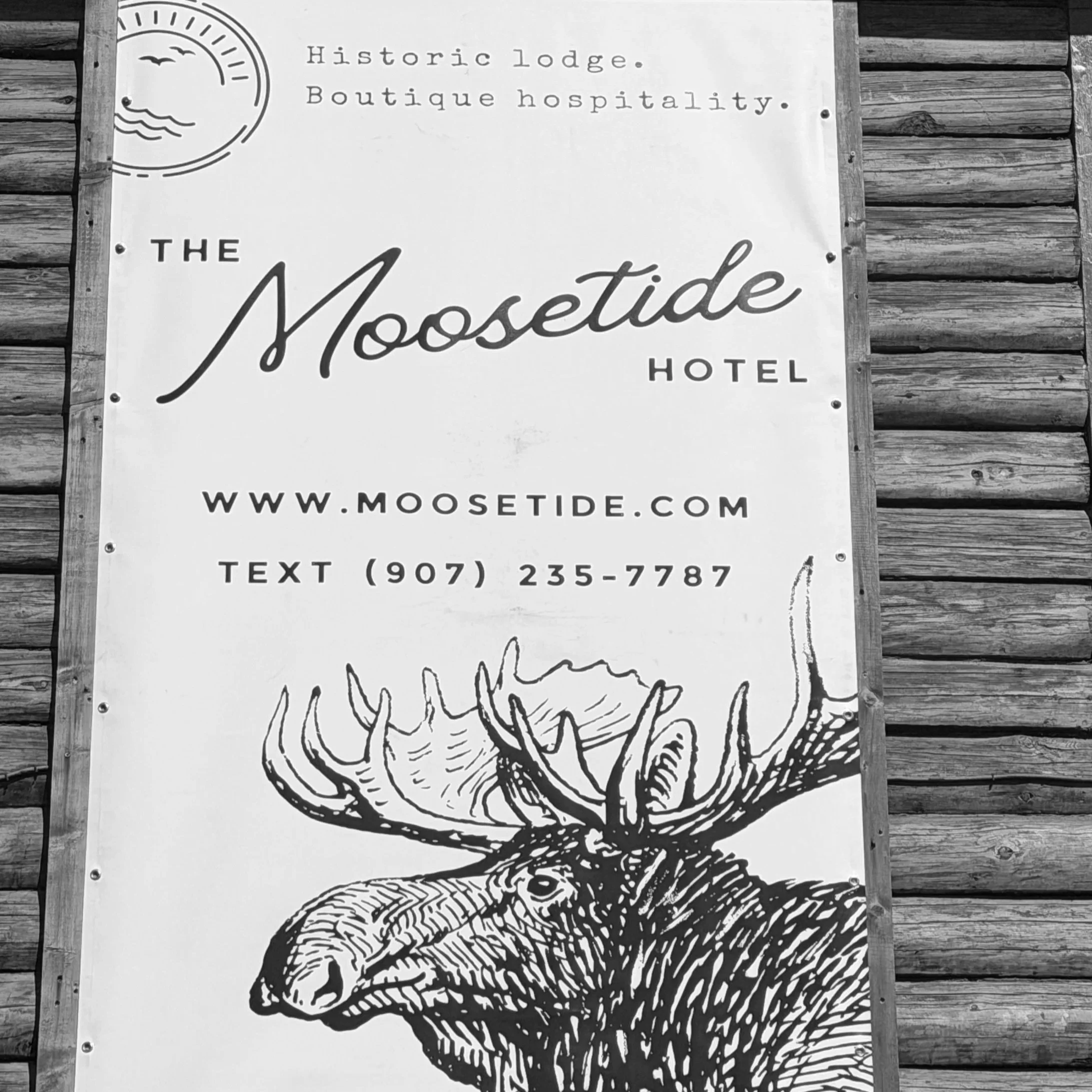 A printed sign on a wooden wall promoting The Moosetide Hotel, featuring a moose illustration and website URL.