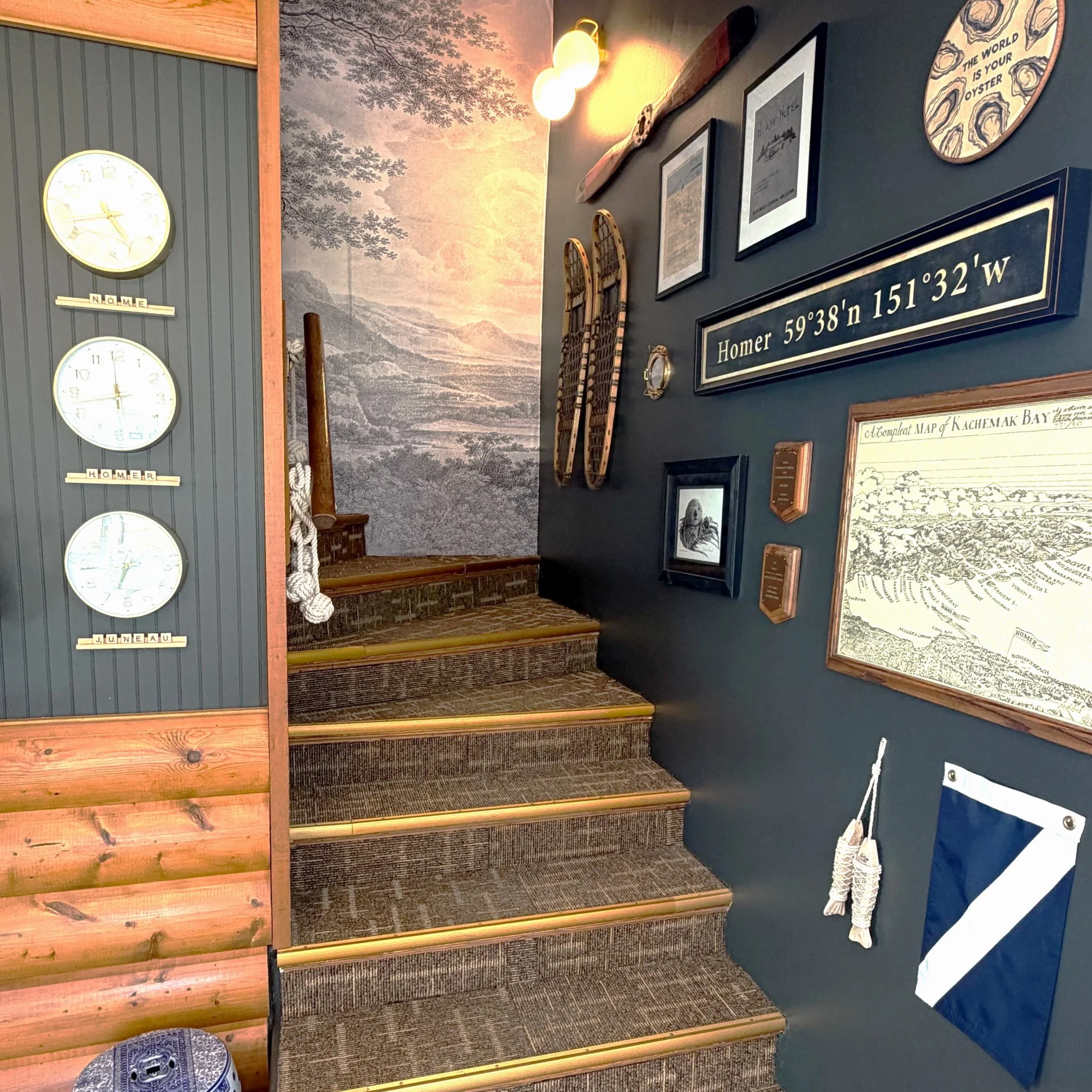 Display of vintage wall clocks, framed photographs, maps, and decorative items on a dark blue wall with a mural of trees and mountains in the background.