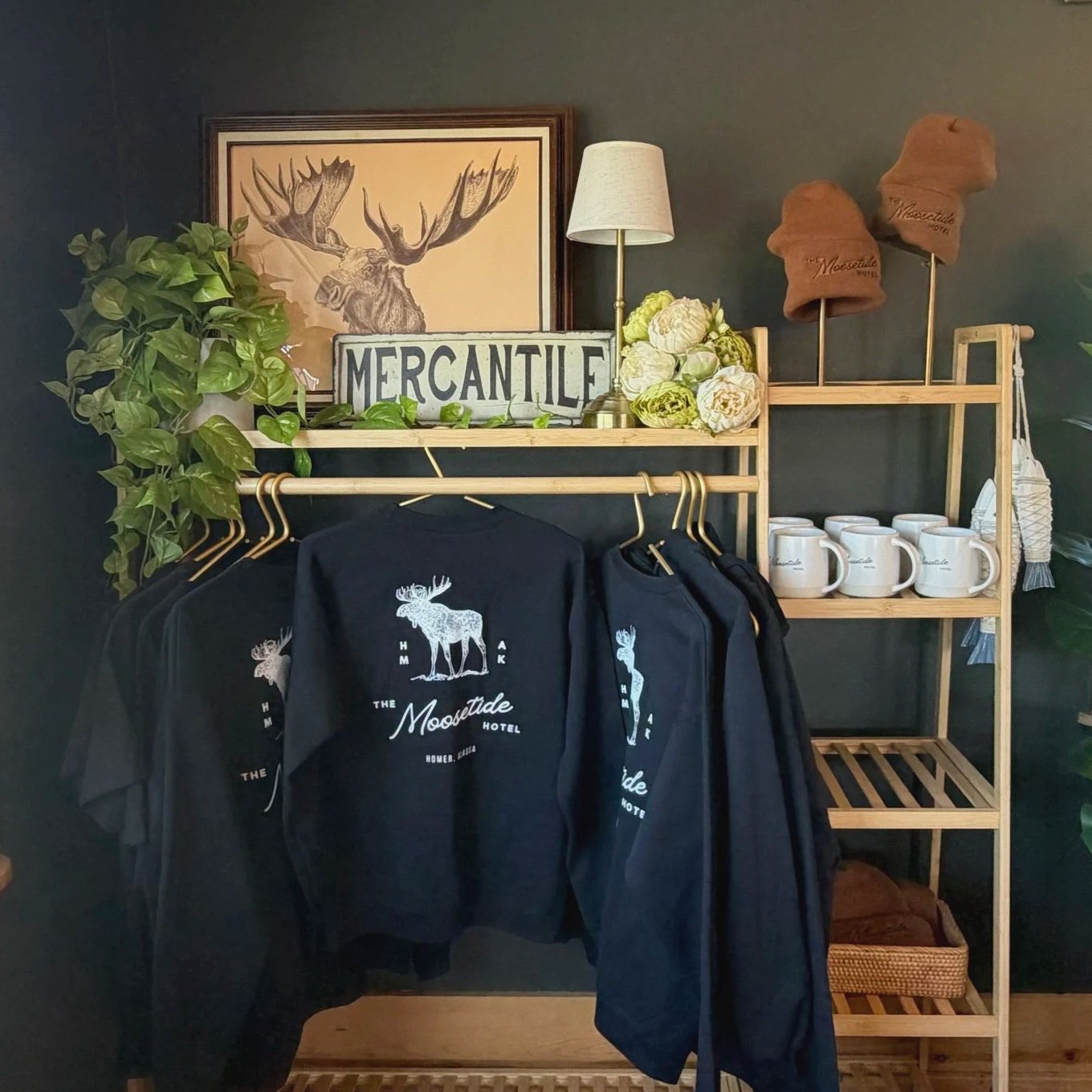 Display shelf with Moose-themed merchandise including a black hoodie with a moose graphic, white mugs, and brown hats. The background includes a framed drawing of a moose and a 'Mercantile' sign, along with plants and a small table.