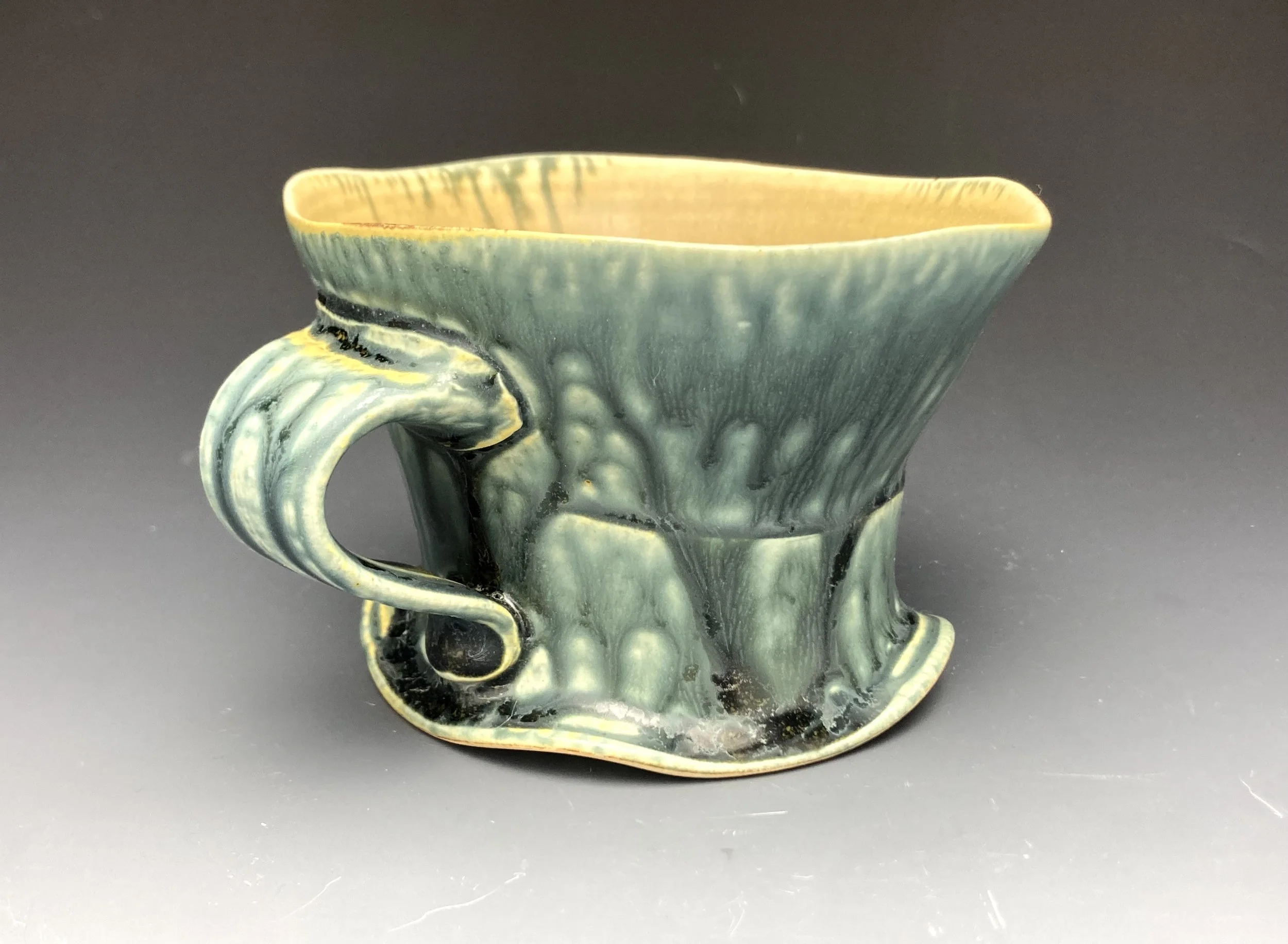 A ceramic mug with green, yellow, and black glaze, featuring a textured, abstract design with a curved handle, set against a dark background.  By Conner Burns