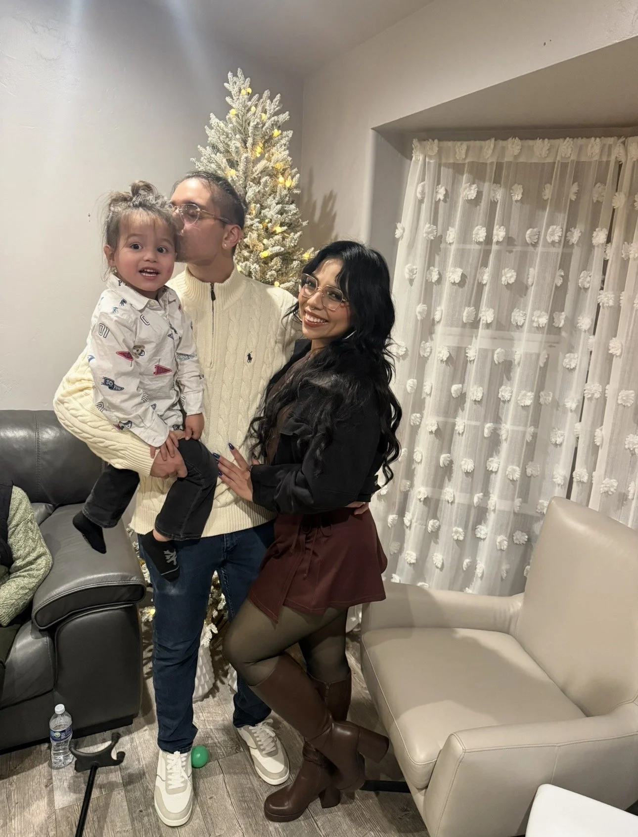 Family celebrating Christmas indoors with a decorated tree, featuring a man, a woman, and a young girl. The man and girl are kissing, and the woman is smiling. There is a couch, a water bottle, and curtains in the background.