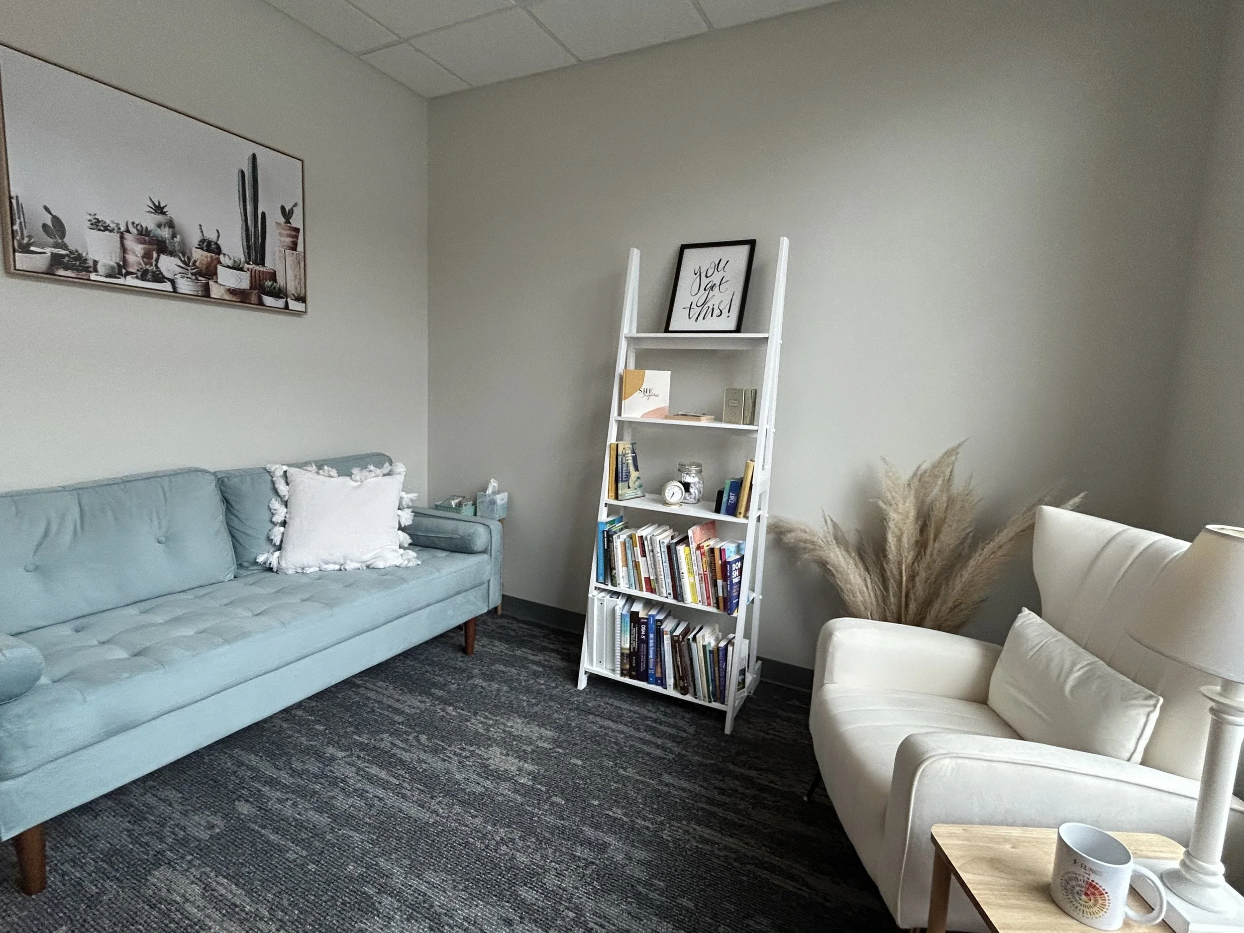 Cozy living room with a light blue sofa, white armchair, bookshelf, artwork, and decorative plants.
