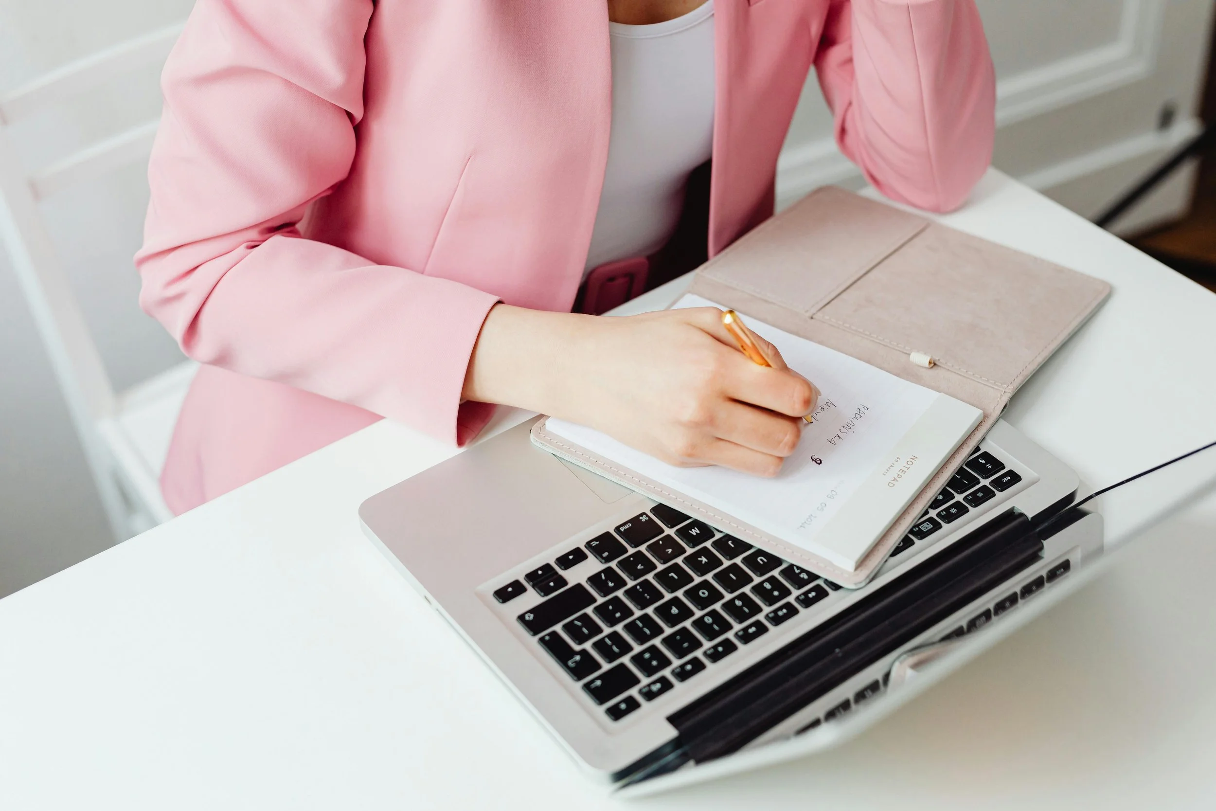 A woman in a pink blazer writing in a notebook on a laptop desk with a closed beige notebook and a laptop.