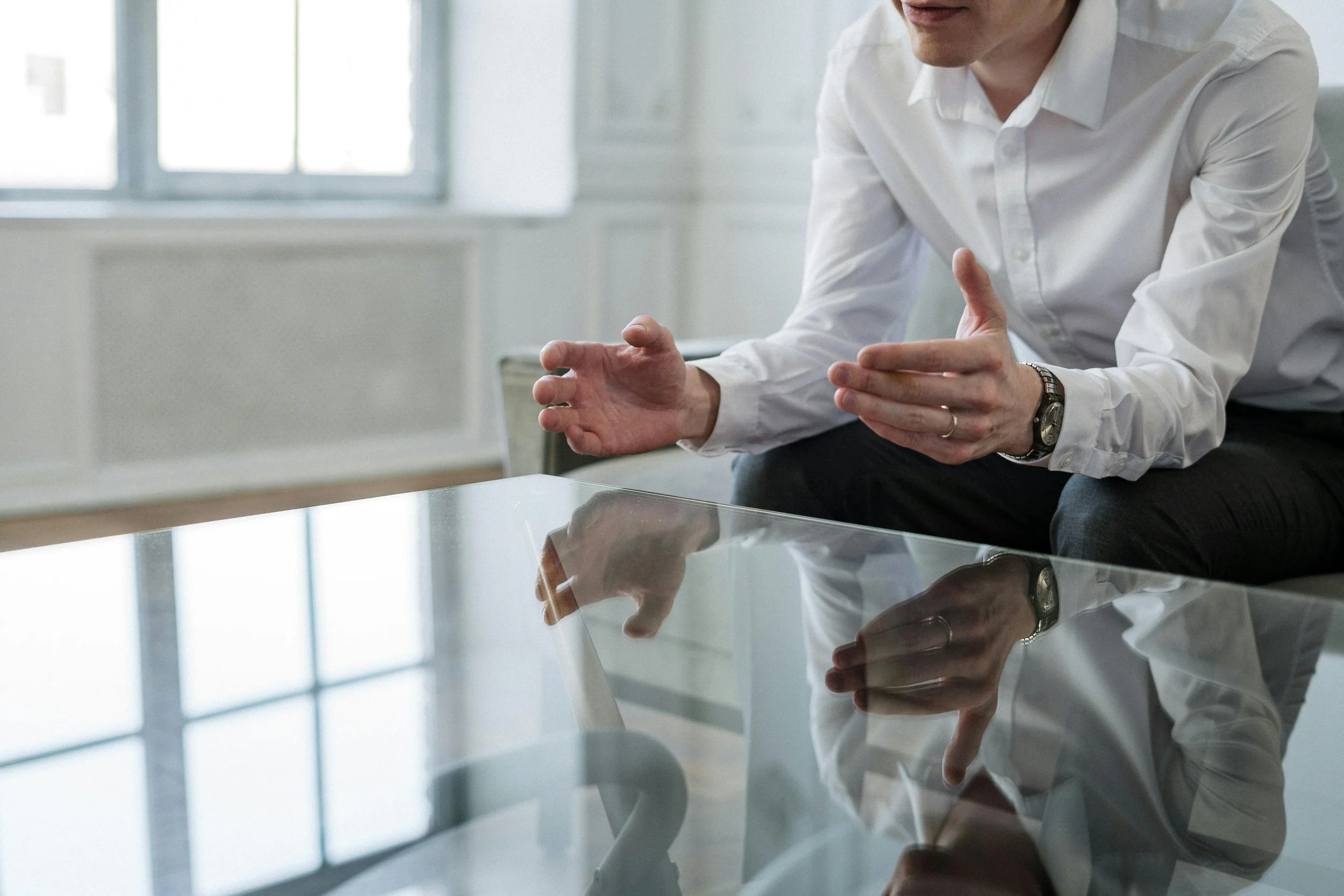 A person in a white shirt with rolled-up sleeves sitting at a glass table, gesturing with hands while speaking, with a reflection of themselves visible on the table surface.