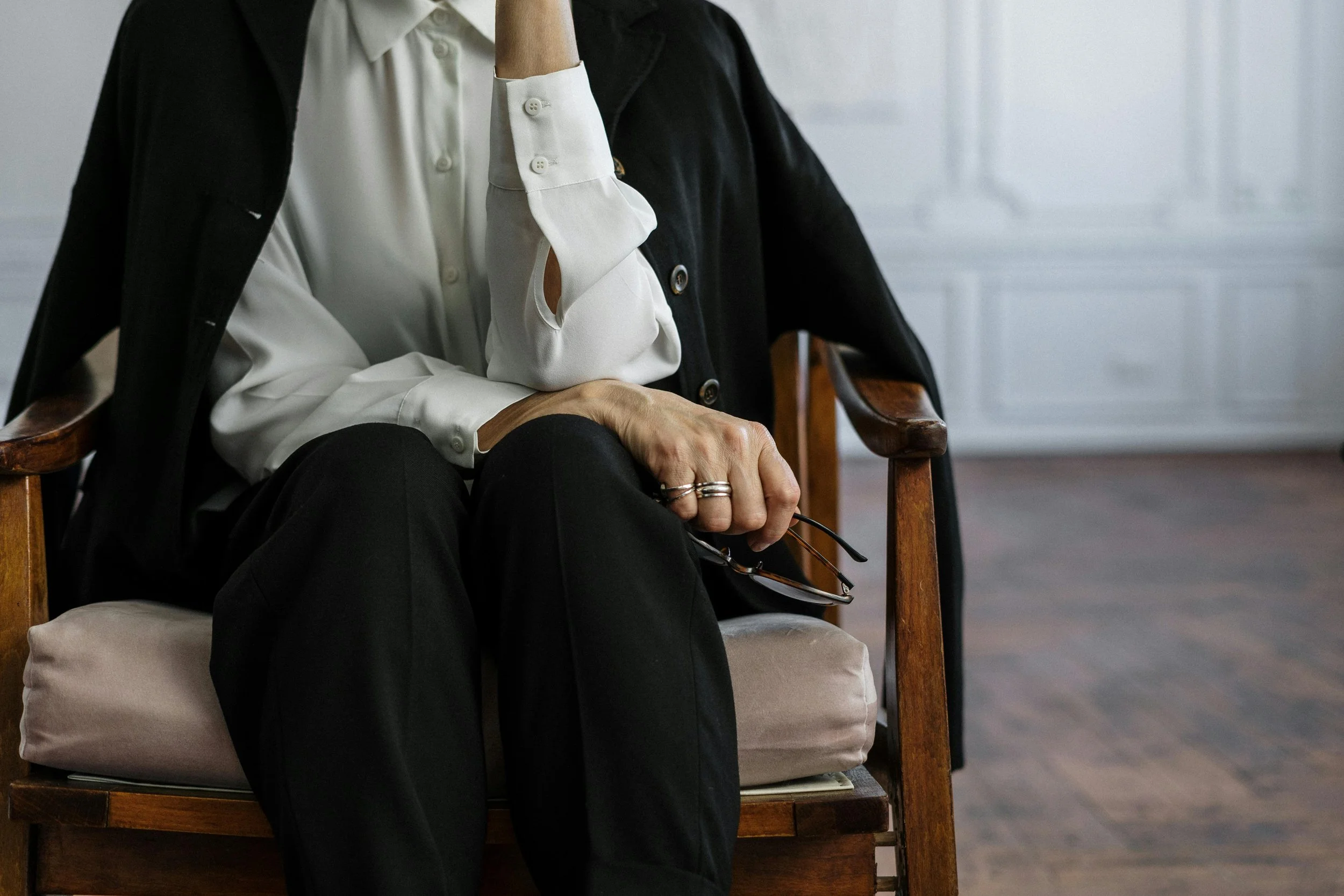 Close-up of a person sitting on a wooden chair with beige cushion, wearing a white shirt, black pants, and a black blazer draped over the shoulders, holding glasses in hand, in a room with white paneled walls and wooden floor.