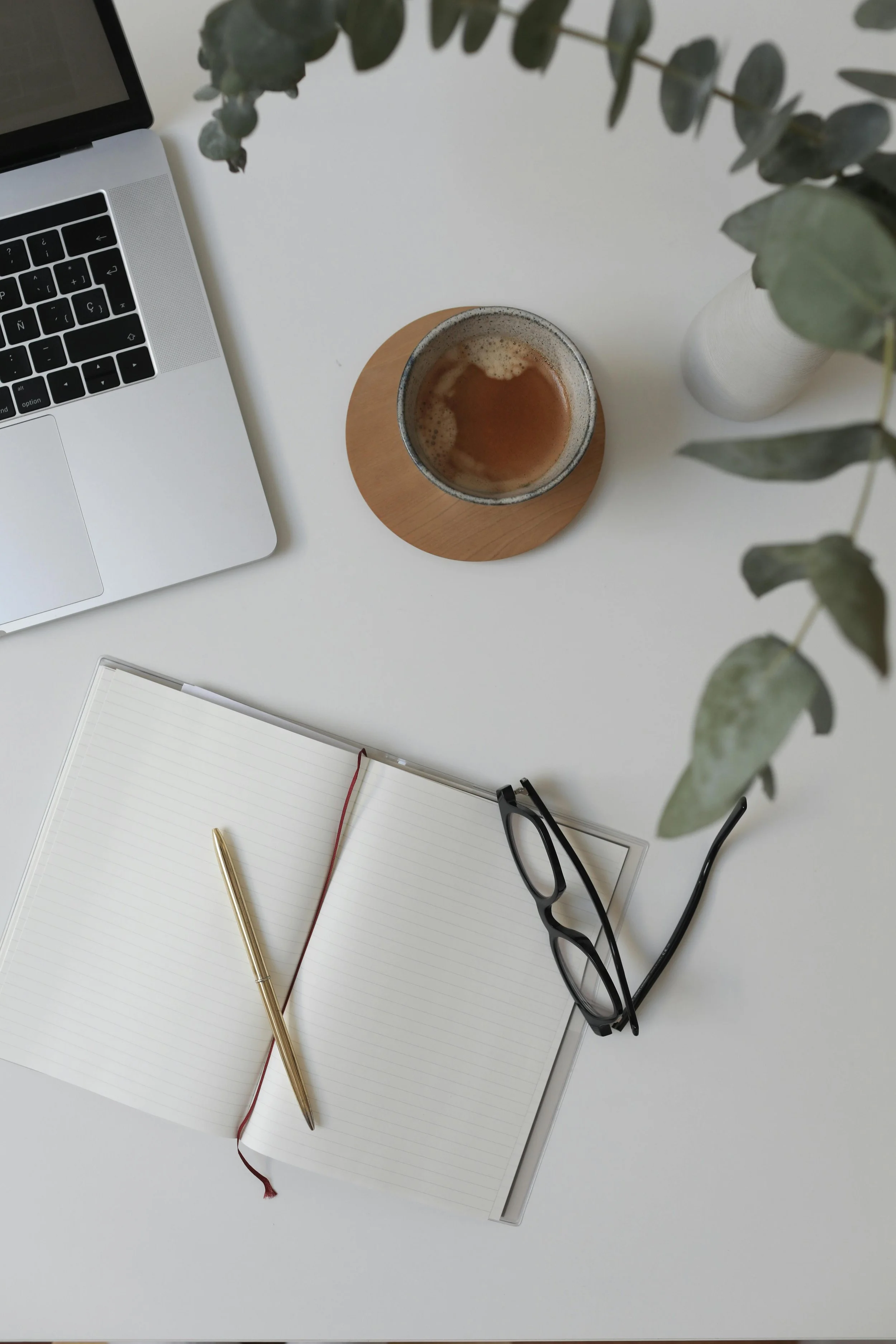 Flat lay of a workspace with a silver laptop, open notebook with a gold pen, black glasses, a coffee cup on a wooden coaster, and a white vase with eucalyptus leaves on a white table.