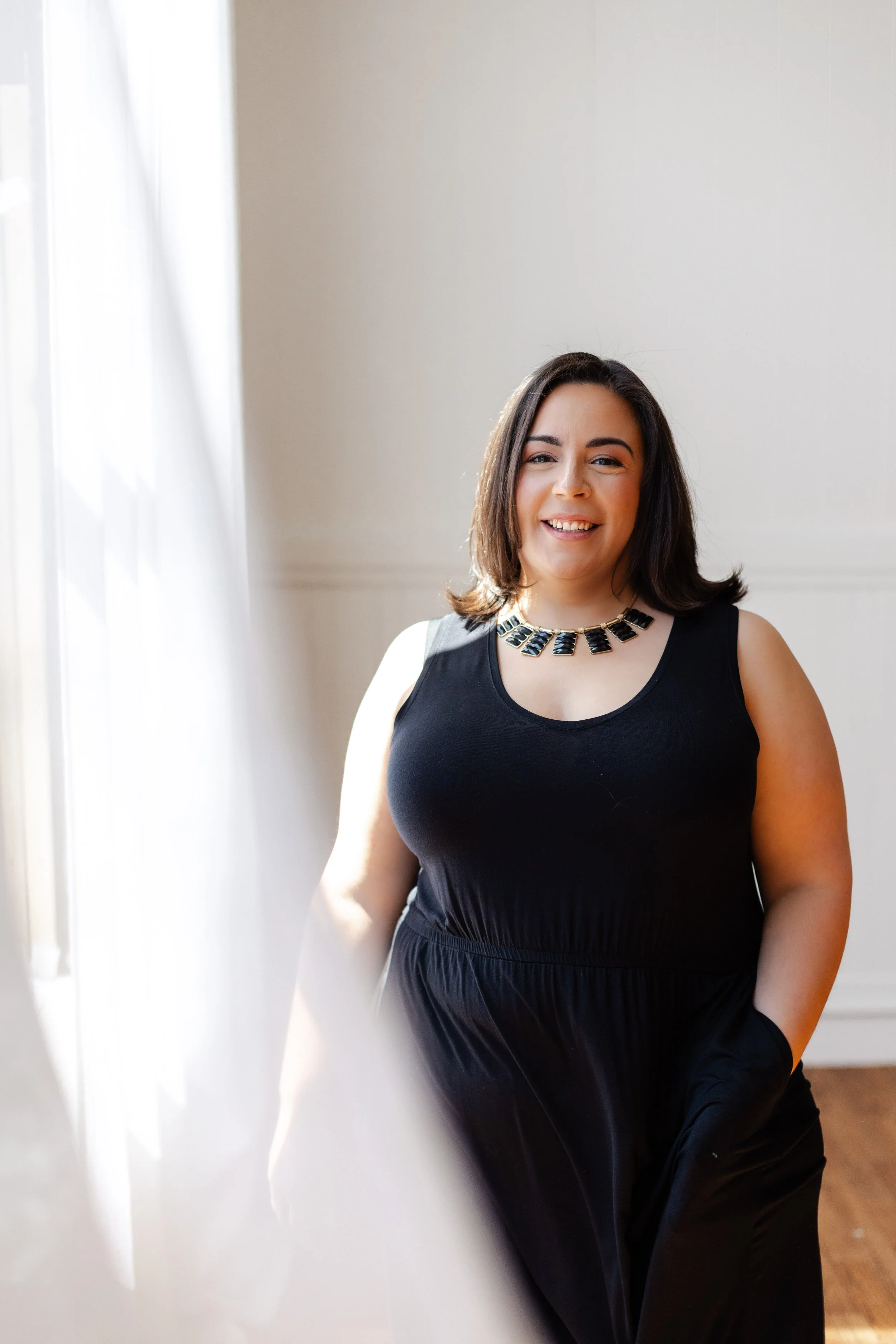 A woman with dark brown hair wearing a black sleeveless dress and a statement necklace, smiling and winking, standing in a room with white walls and sunlight streaming through a window.