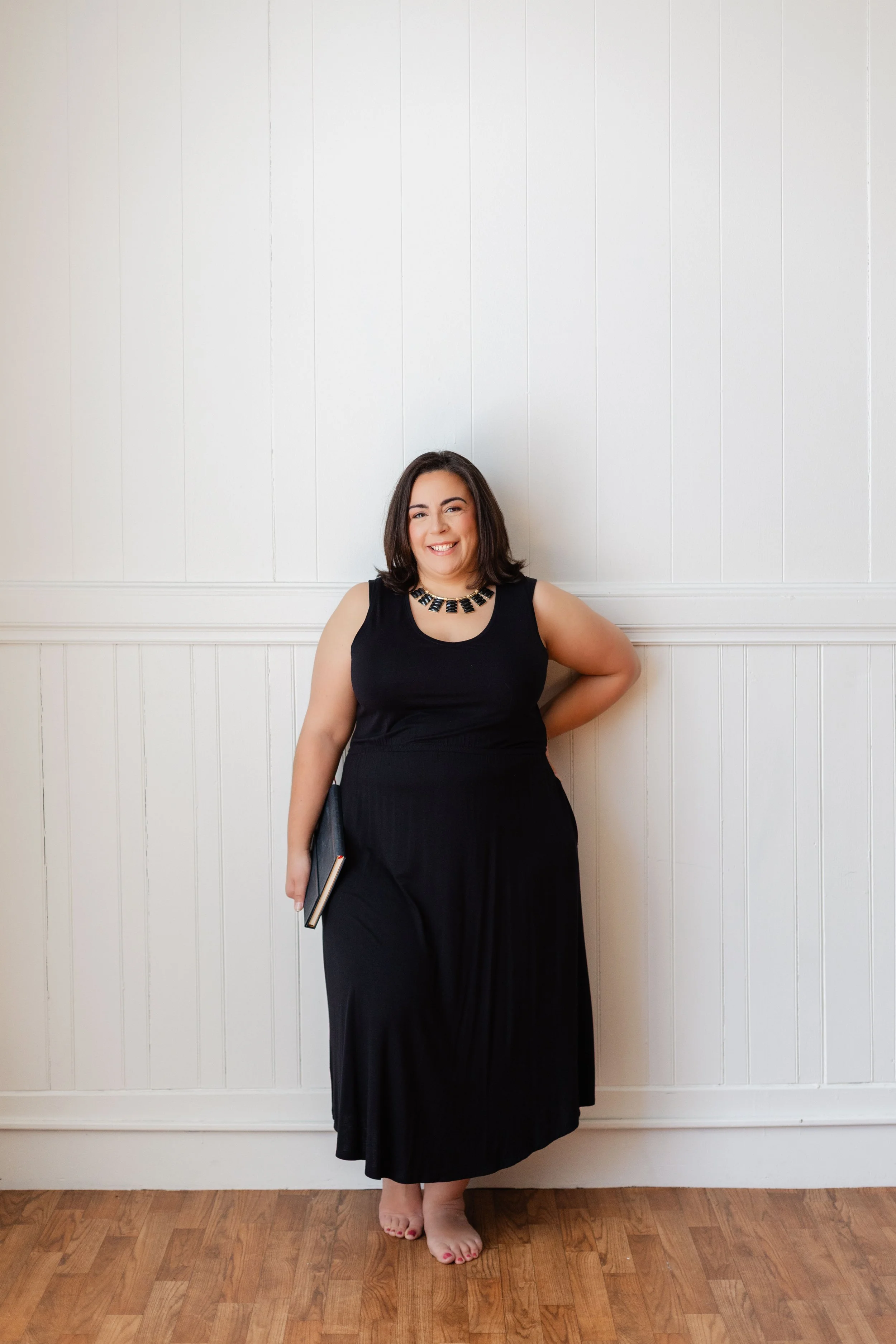 A woman dressed in a black sleeveless top and a long black skirt, standing barefoot against a white paneled wall, holding a book in her right hand, smiling and looking at the camera.