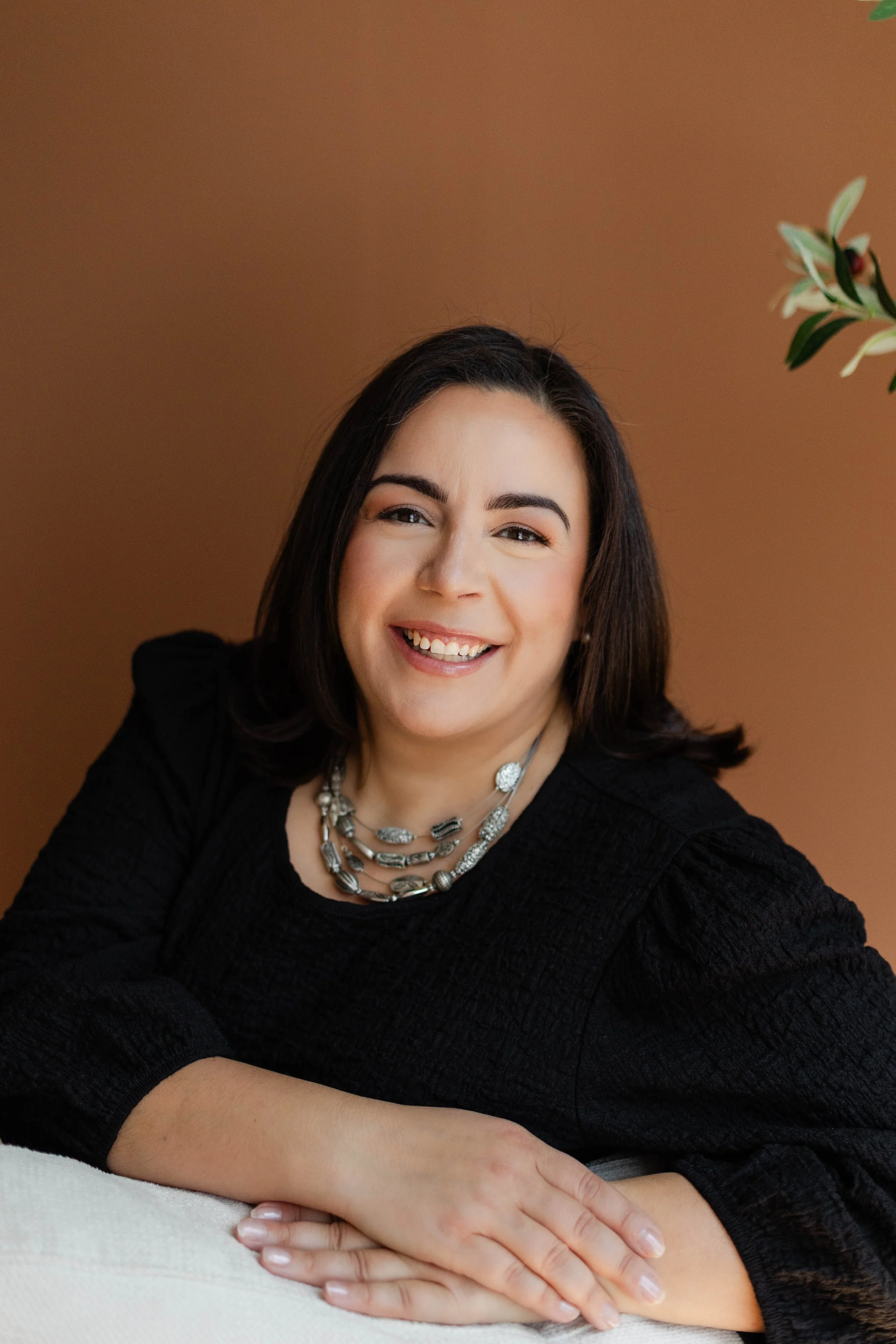 A woman with dark hair smiling, wearing a black top and layered silver necklaces, sitting with her arms on a white surface against a brown background.