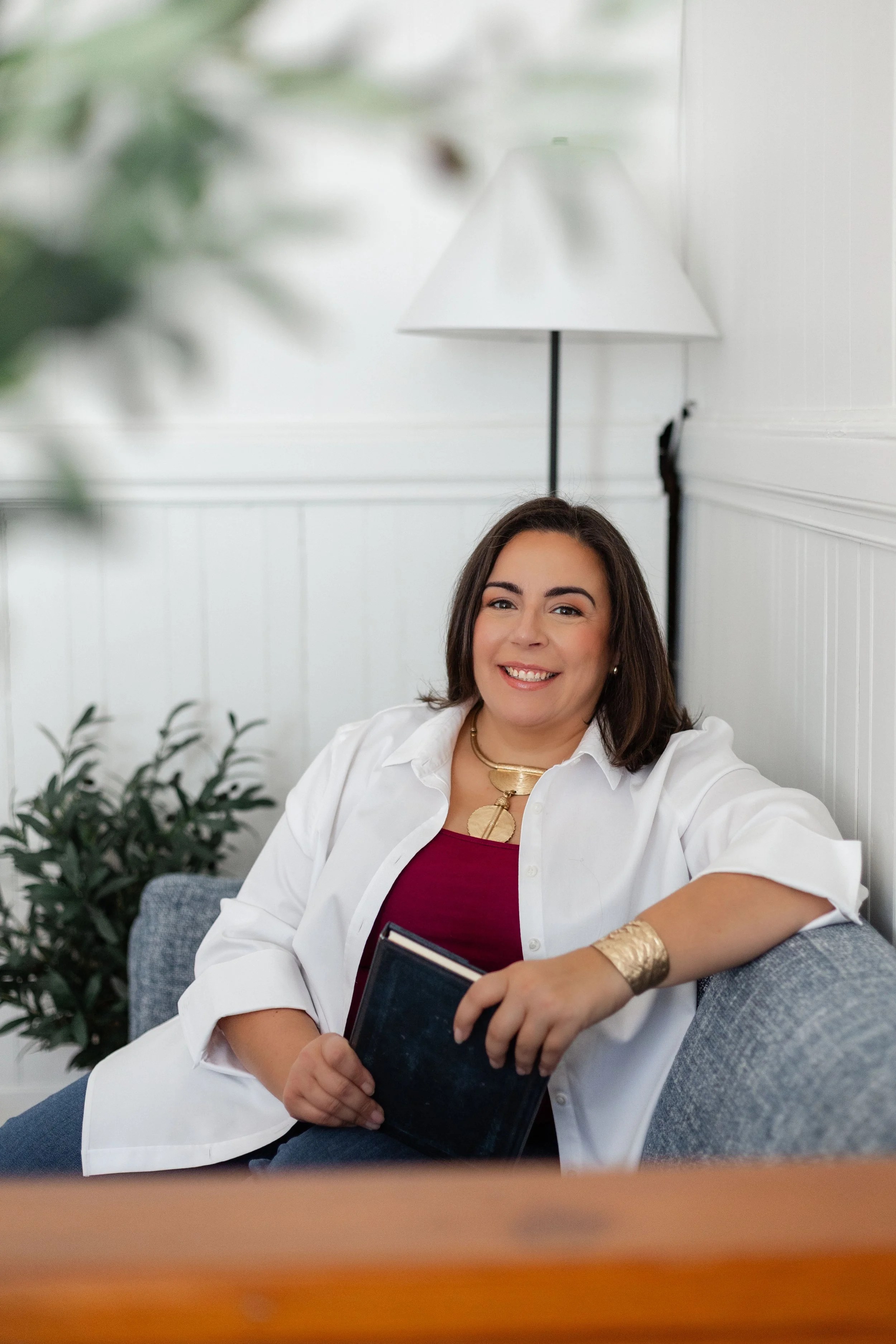 A woman with shoulder-length brown hair, wearing a white shirt, red top, gold necklace, and bracelet, sitting on a gray couch in a modern room with white paneled walls and a lamp in the background, smiling and holding a tablet.