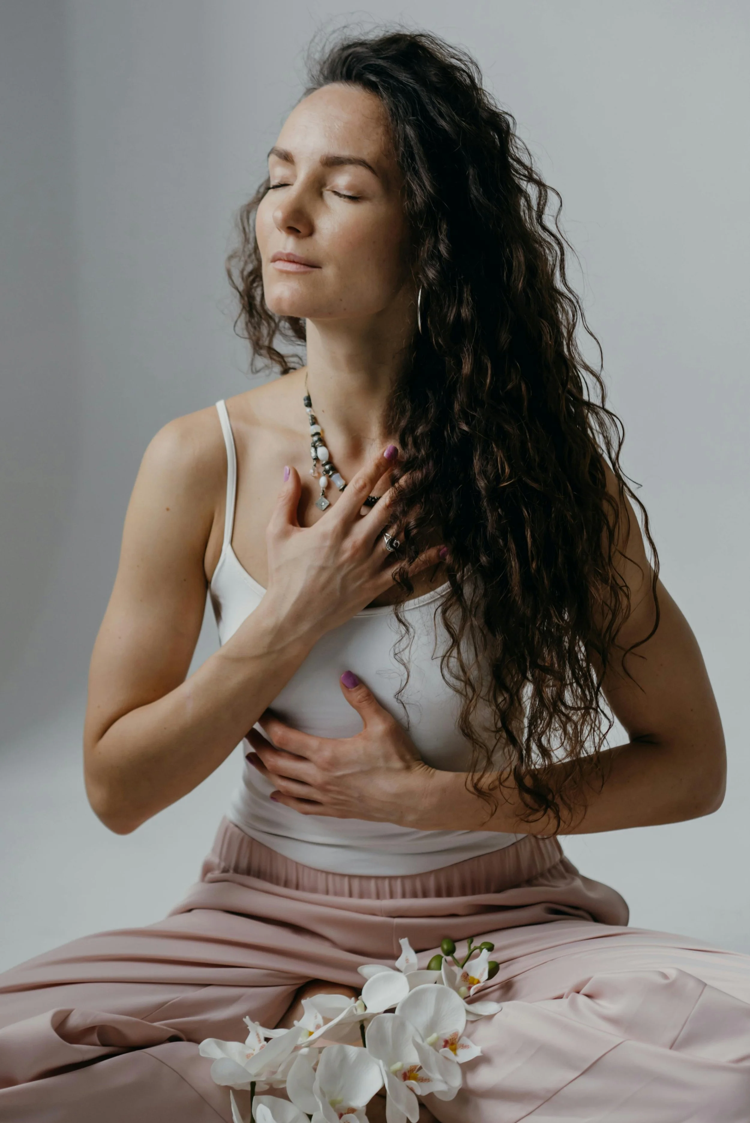 A woman with long, curly hair sitting with her eyes closed, hands over her chest and stomach, wearing a white tank top and beige pants, surrounded by white orchid flowers.