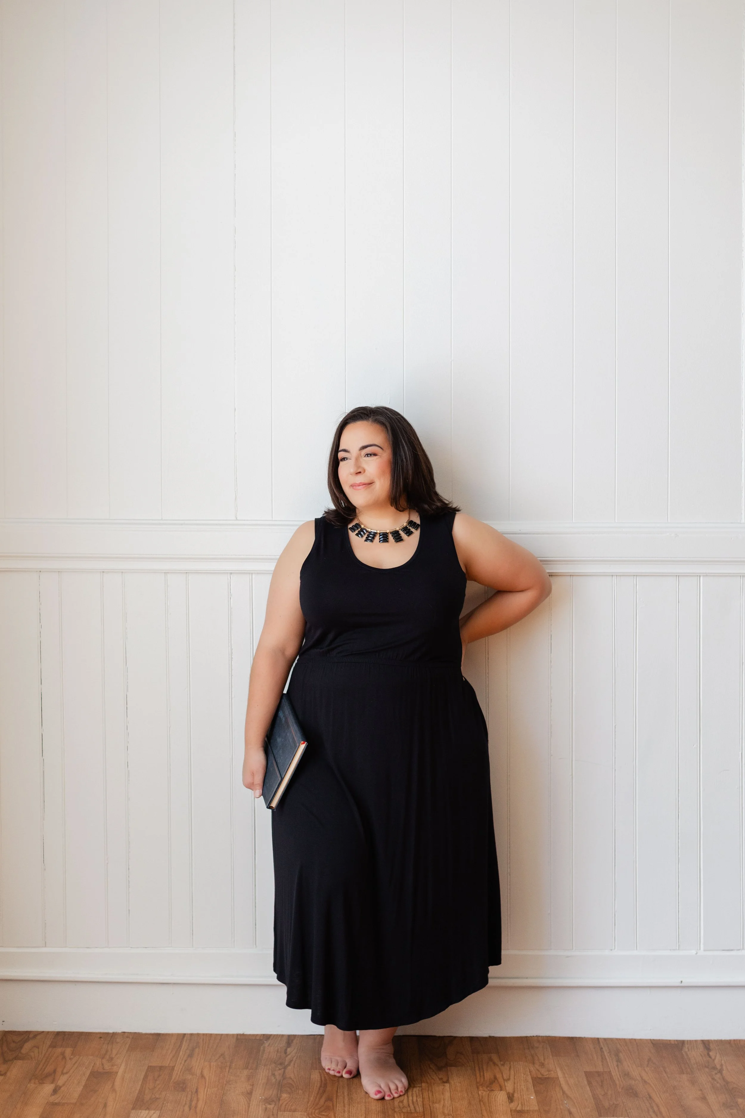 A woman in a black dress with a statement necklace holding a closed book, standing barefoot against a white paneled wall with wooden floors.