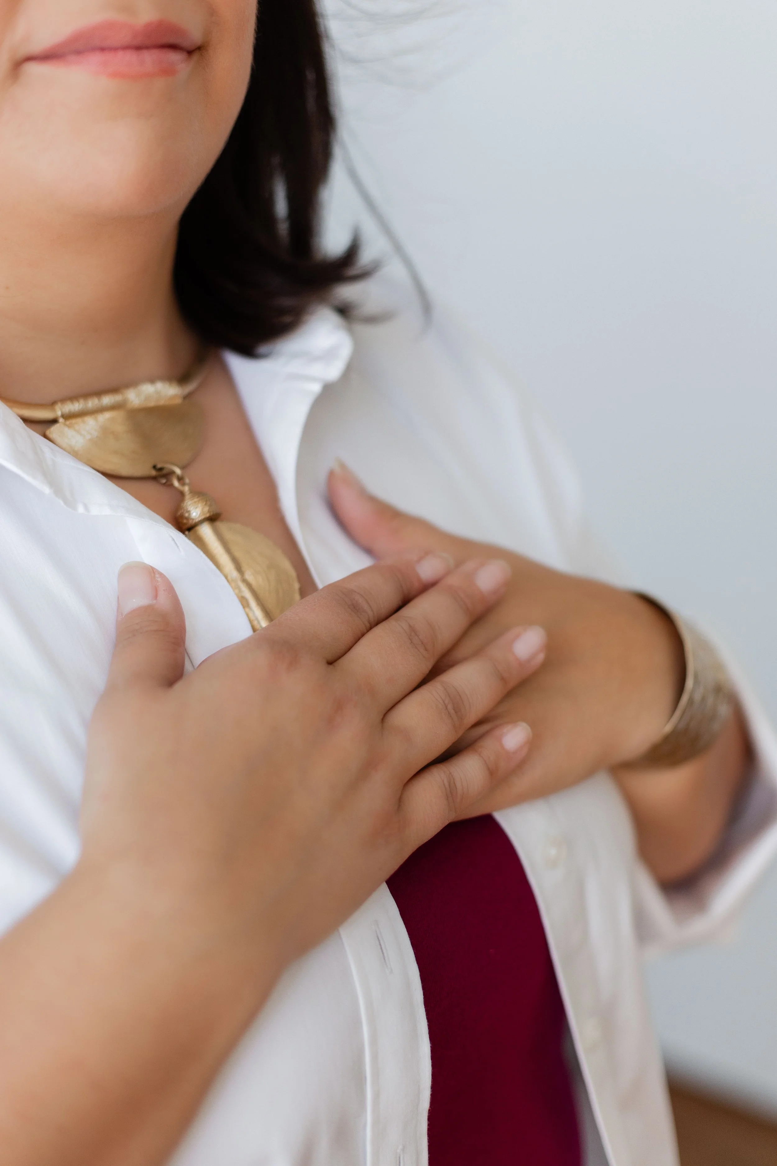 A woman with dark hair and light skin is touching her chest near her collarbone with her left hand. She is wearing a white blouse, a maroon top underneath, and a gold necklace. The background is white.