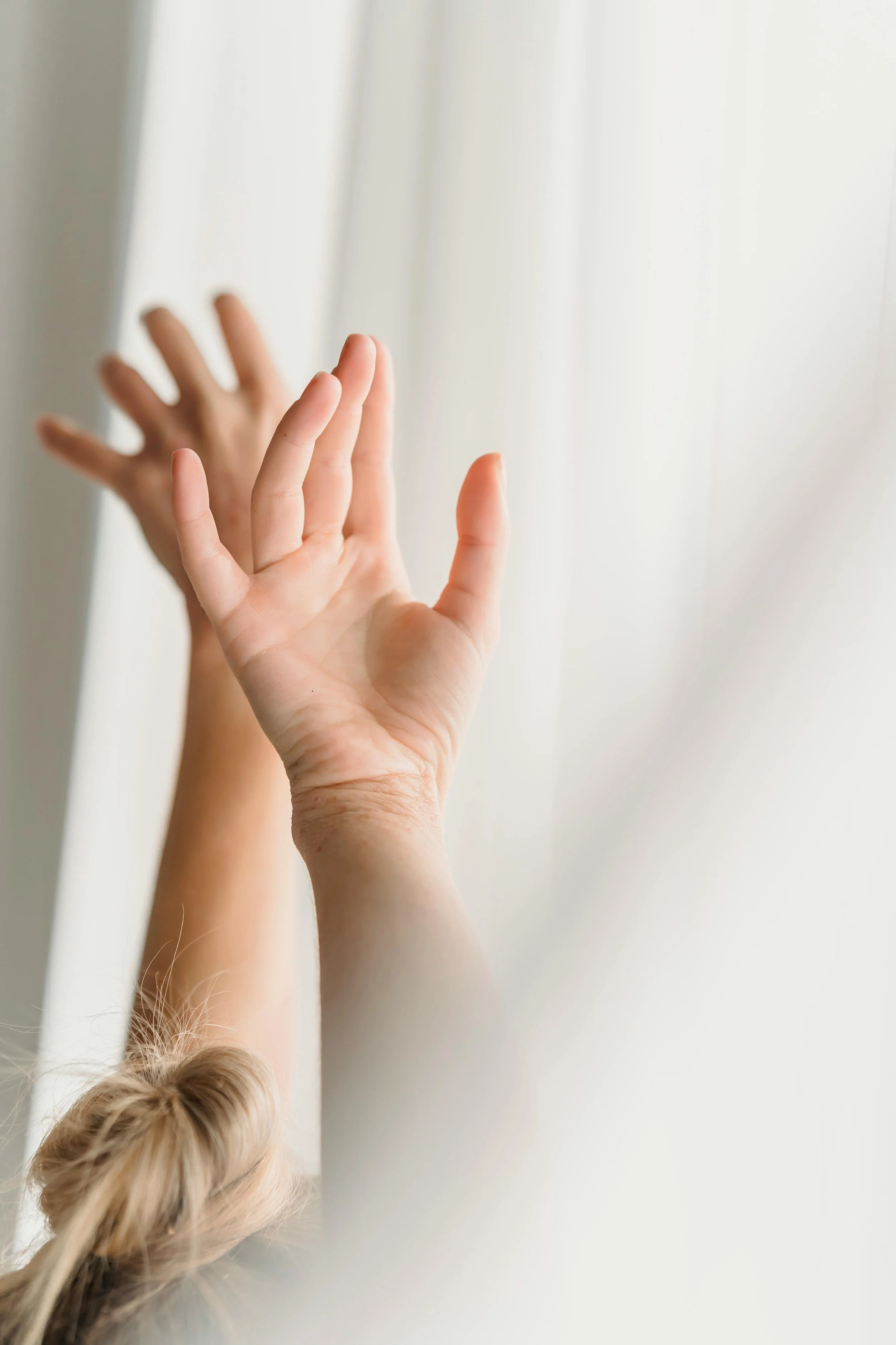 A child's hand reaching out to touch a glass surface, with their reflection visible on the other side, against a light-colored background.