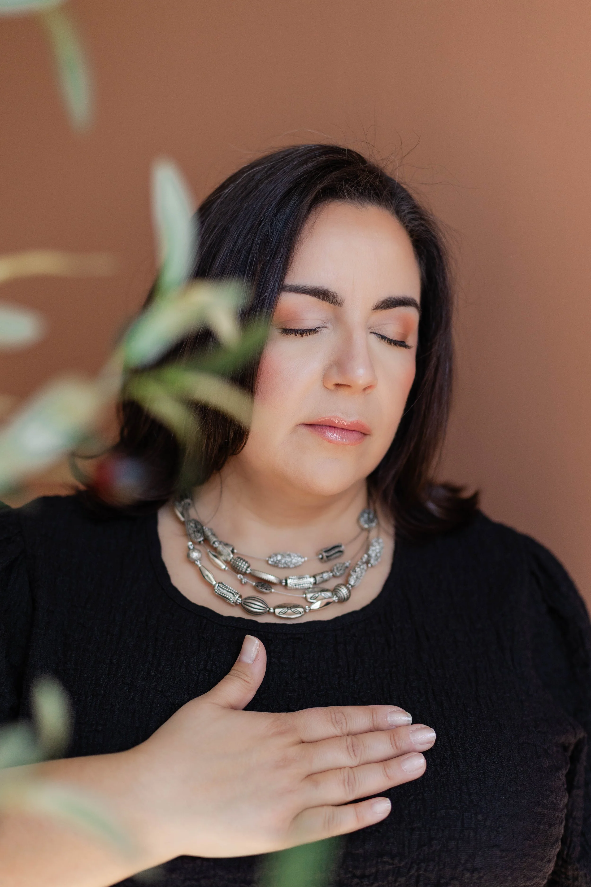 A woman with dark hair and a peaceful expression, eyes closed, wearing layered silver necklaces and a black top. Her right hand is placed on her chest, and her left hand is near her face, with a blurred green plant in the foreground and a warm brown background.