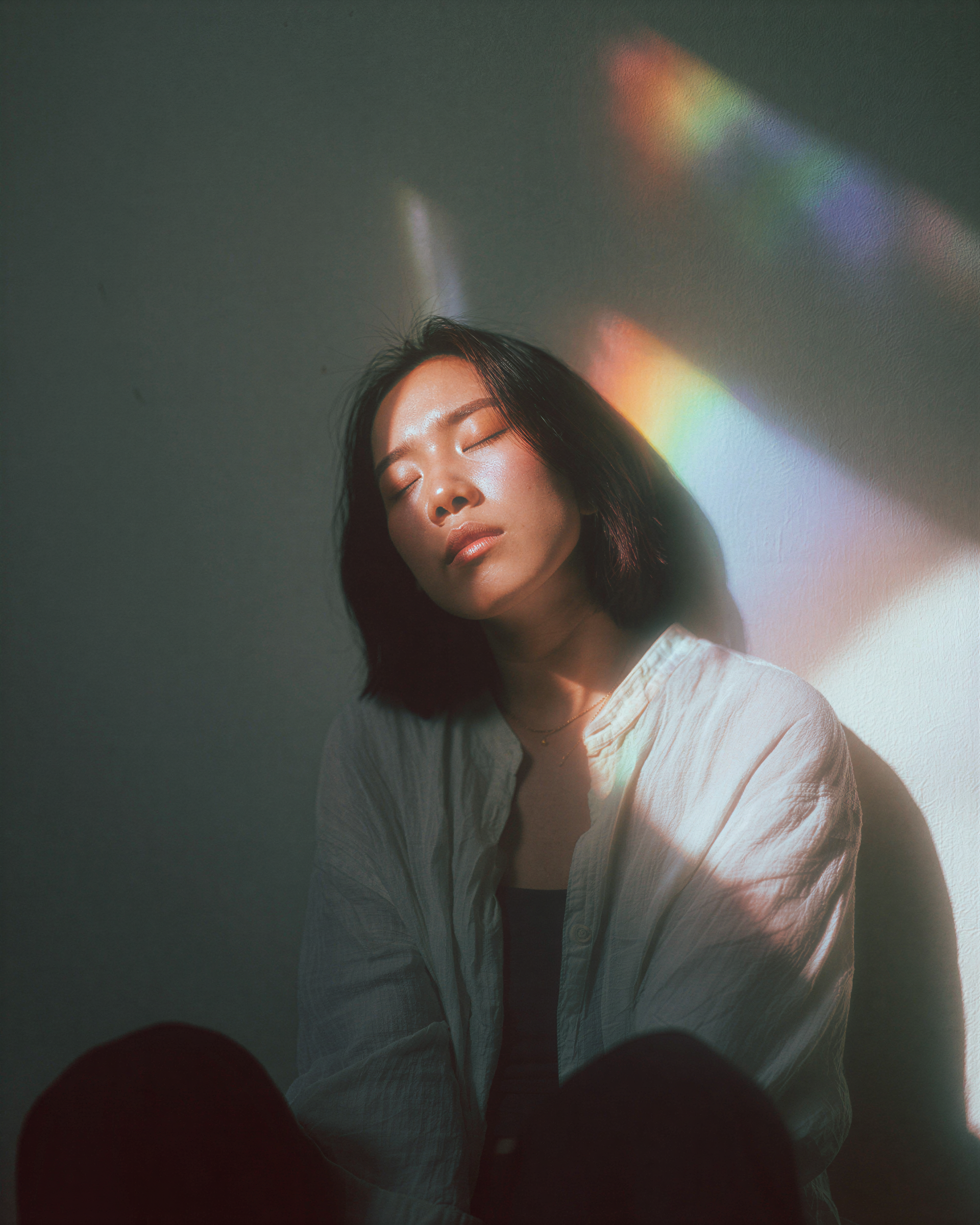 A woman with medium skin tone and dark hair, eyes closed, standing or sitting against a plain wall with rainbow-colored light reflections on her face and shoulder.