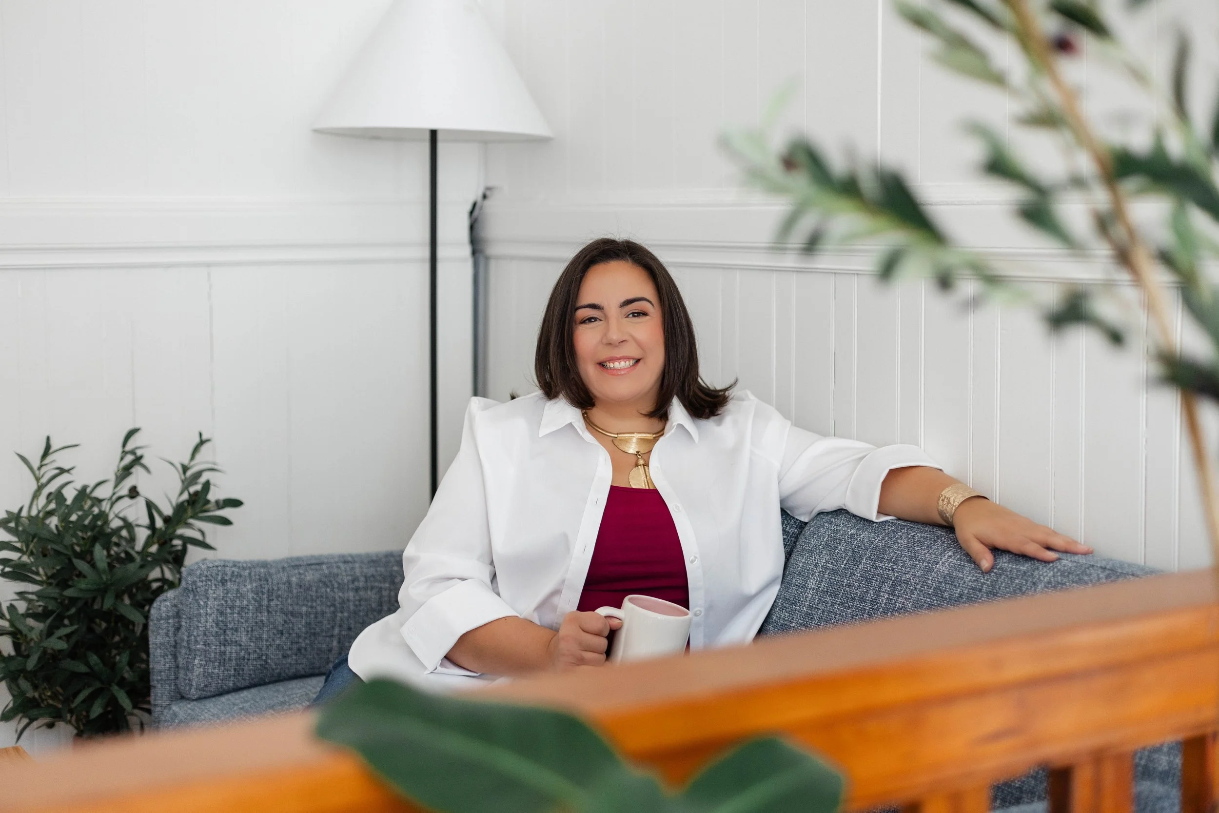 A smiling woman with shoulder-length dark hair, wearing a white shirt over a maroon top, sitting on a gray couch in a bright room with white walls and plants. She is holding a white mug in her right hand.