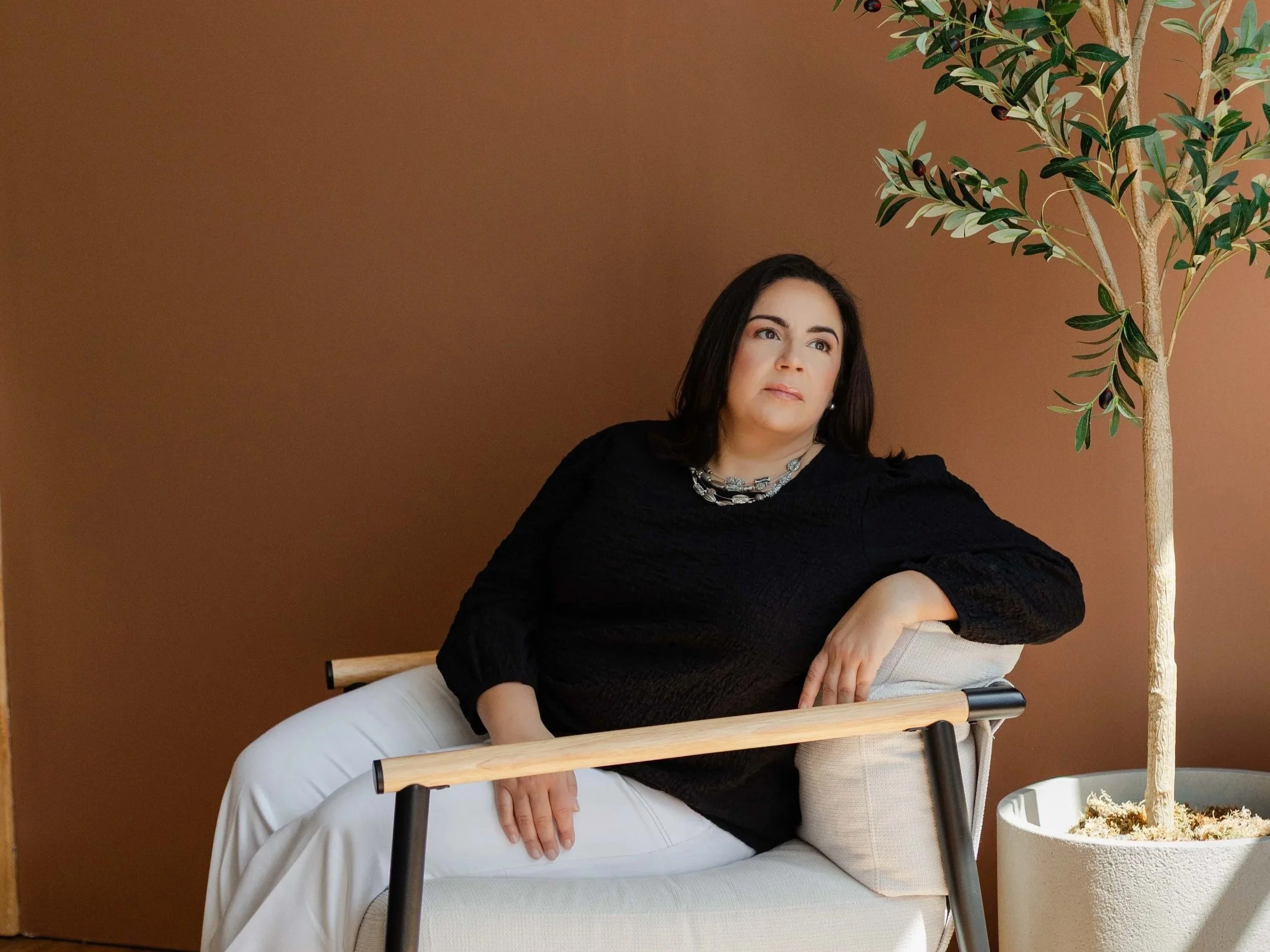 A woman with dark hair wearing a black top and white pants sitting on a chair against a brown background, beside a potted tree with green leaves and dark berries.