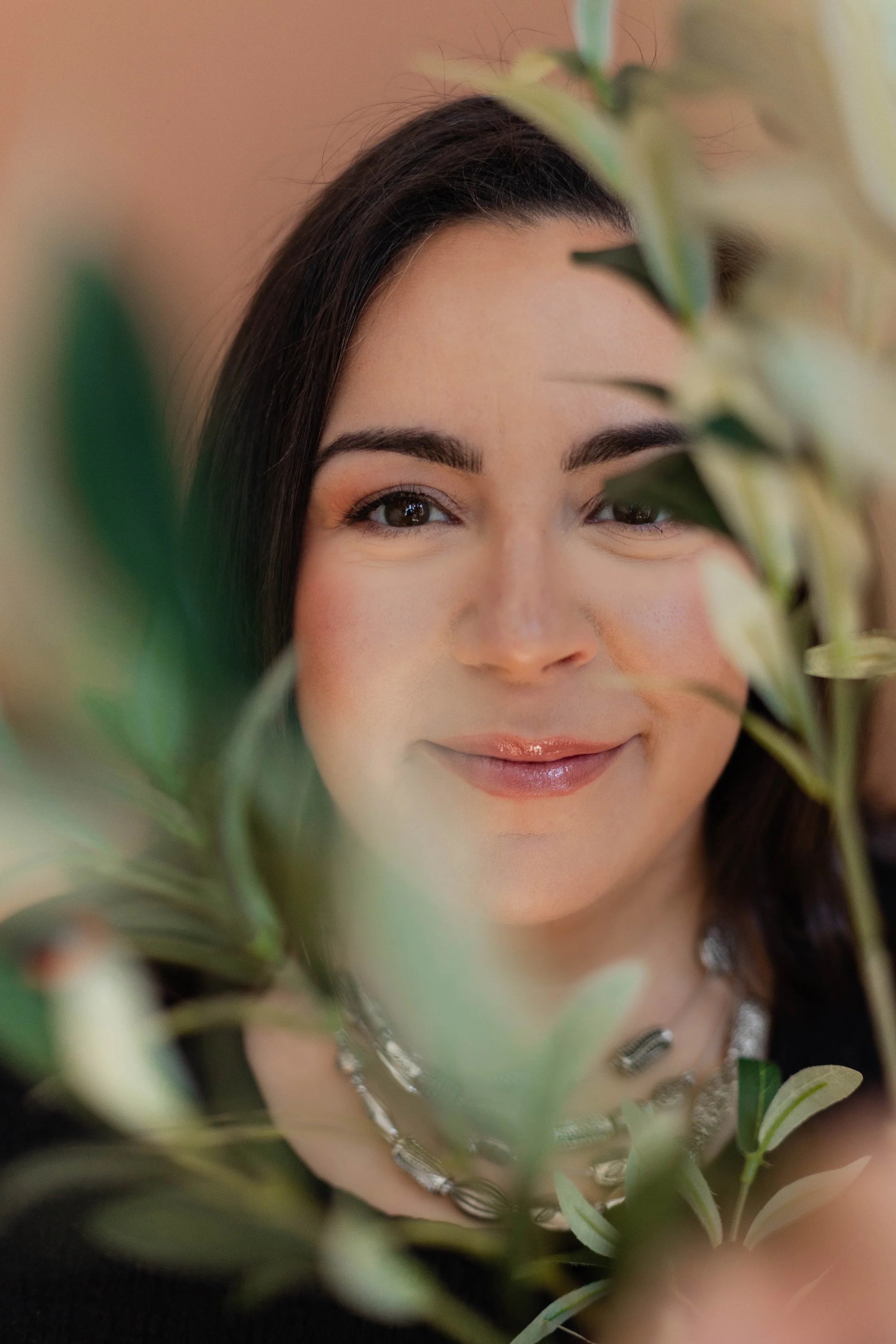 Close-up of a woman with dark hair, makeup, and jewelry, partially obscured by green foliage in the foreground.