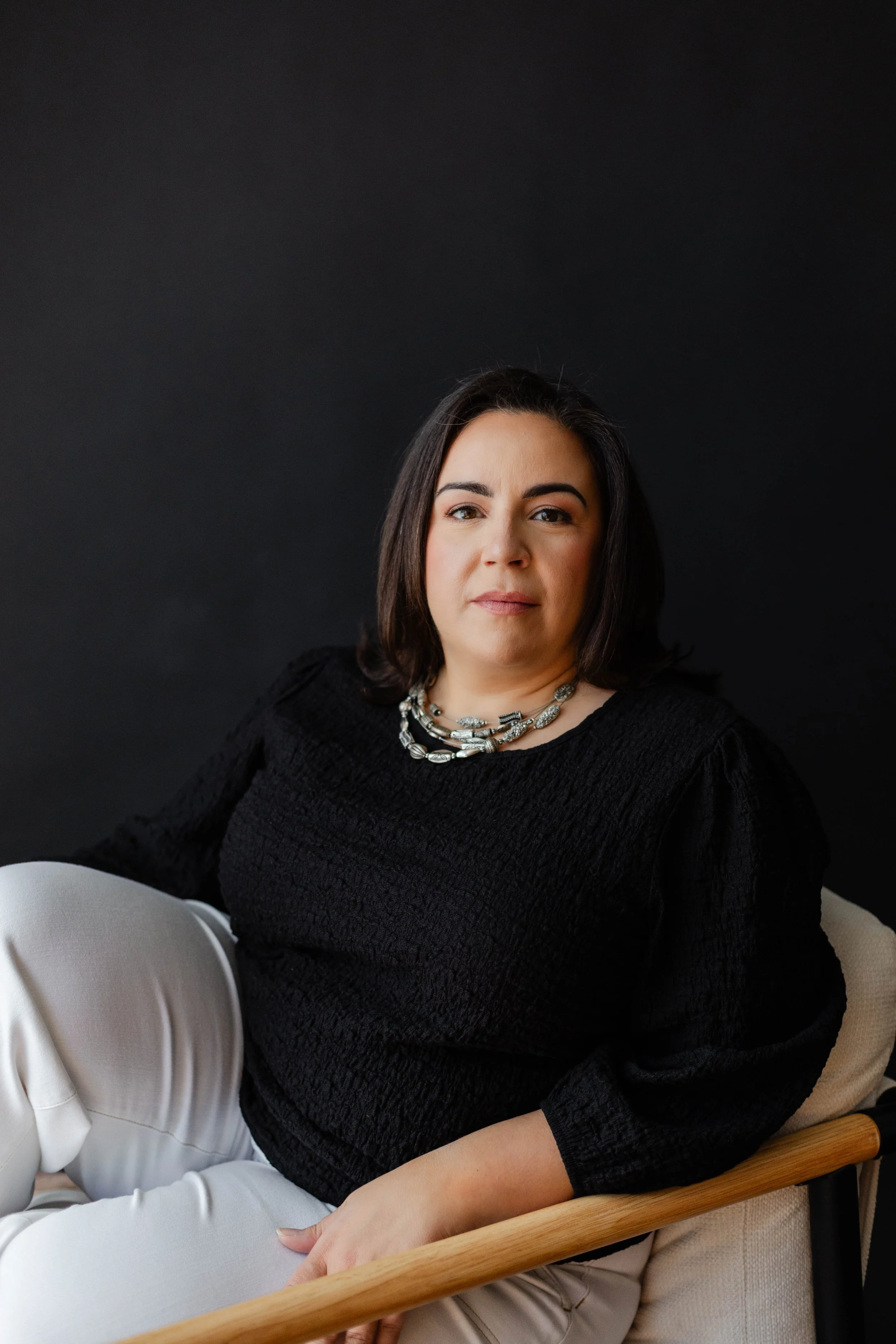 A woman with shoulder-length dark hair wearing a black textured top and a silver statement necklace, sitting on a beige chair against a dark background, with part of a person in white pants visible in the foreground.