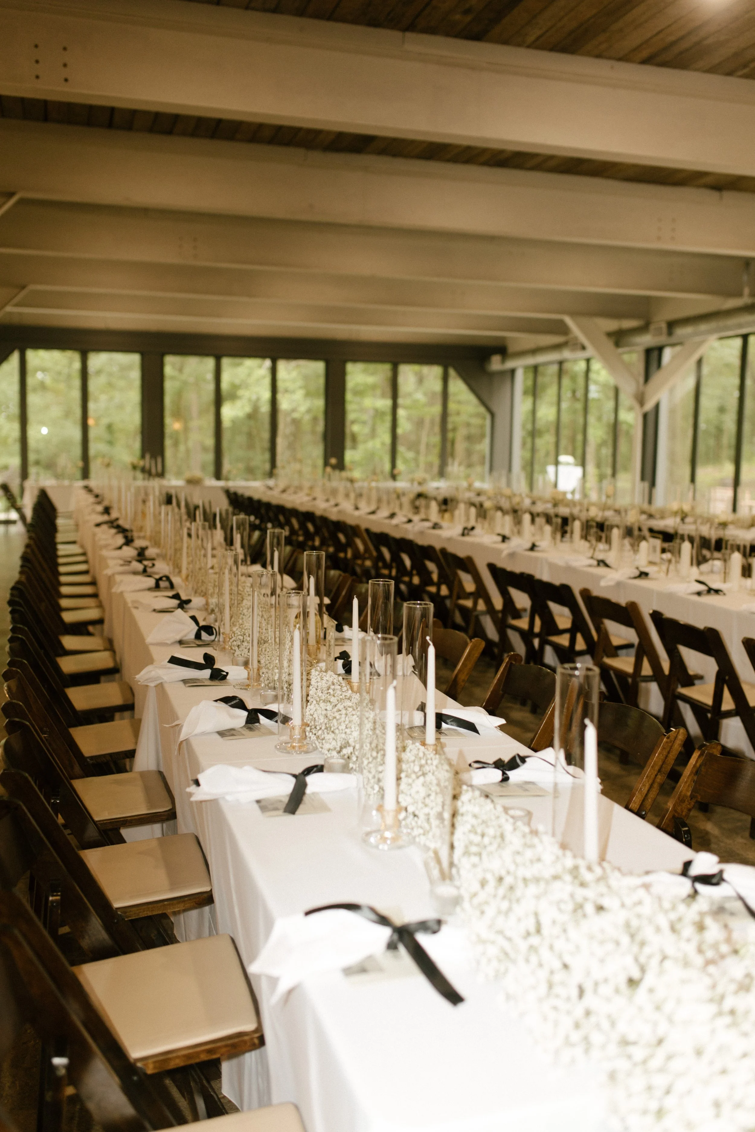 Wedding reception setup with long tables, white tablecloths, white flowers, tall candles, and black ribbons in a room with large windows and wooden ceiling.