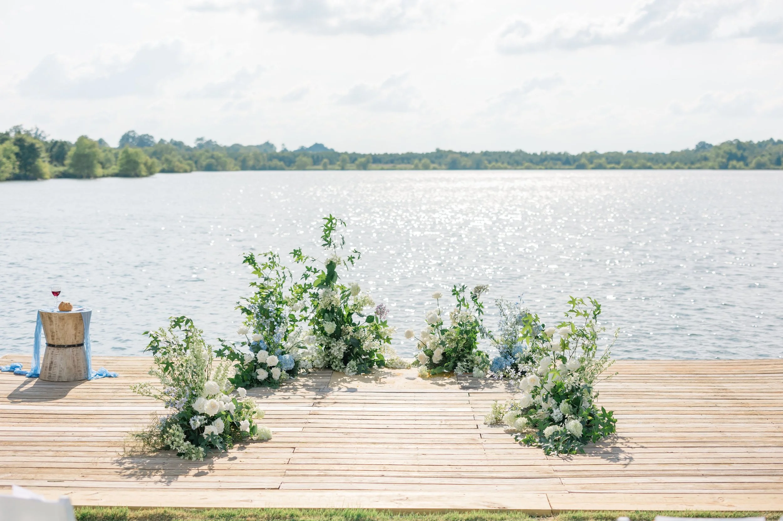 Wedding ceremony setup on a wooden dock by a lake with floral arrangements and a small table with a glass of wine and bread.