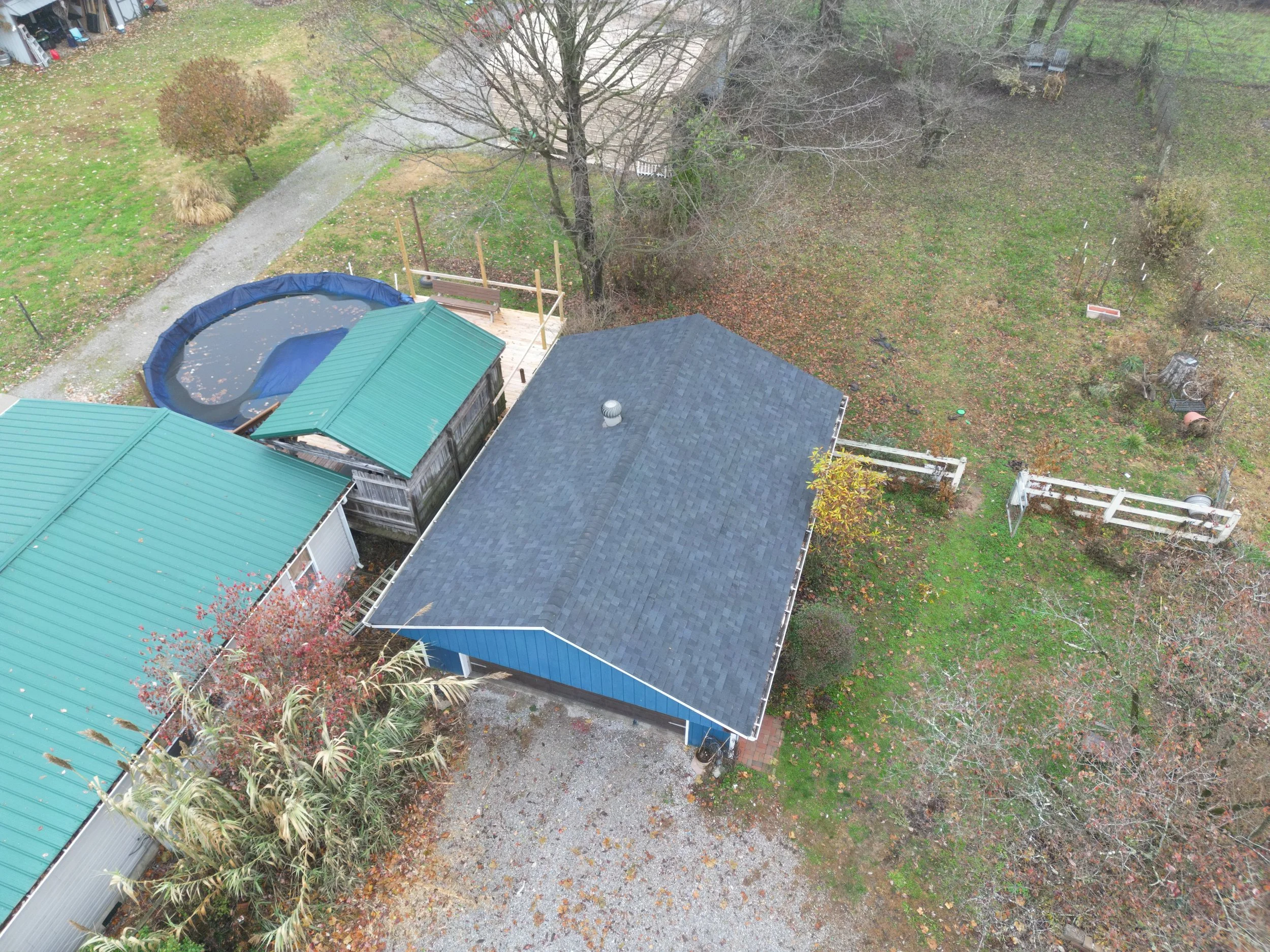 An aerial view of a backyard with a gray roof shed, a green roof shed, and a trampoline with a safety net, surrounded by trees, fences, and grassy areas with fallen leaves.
