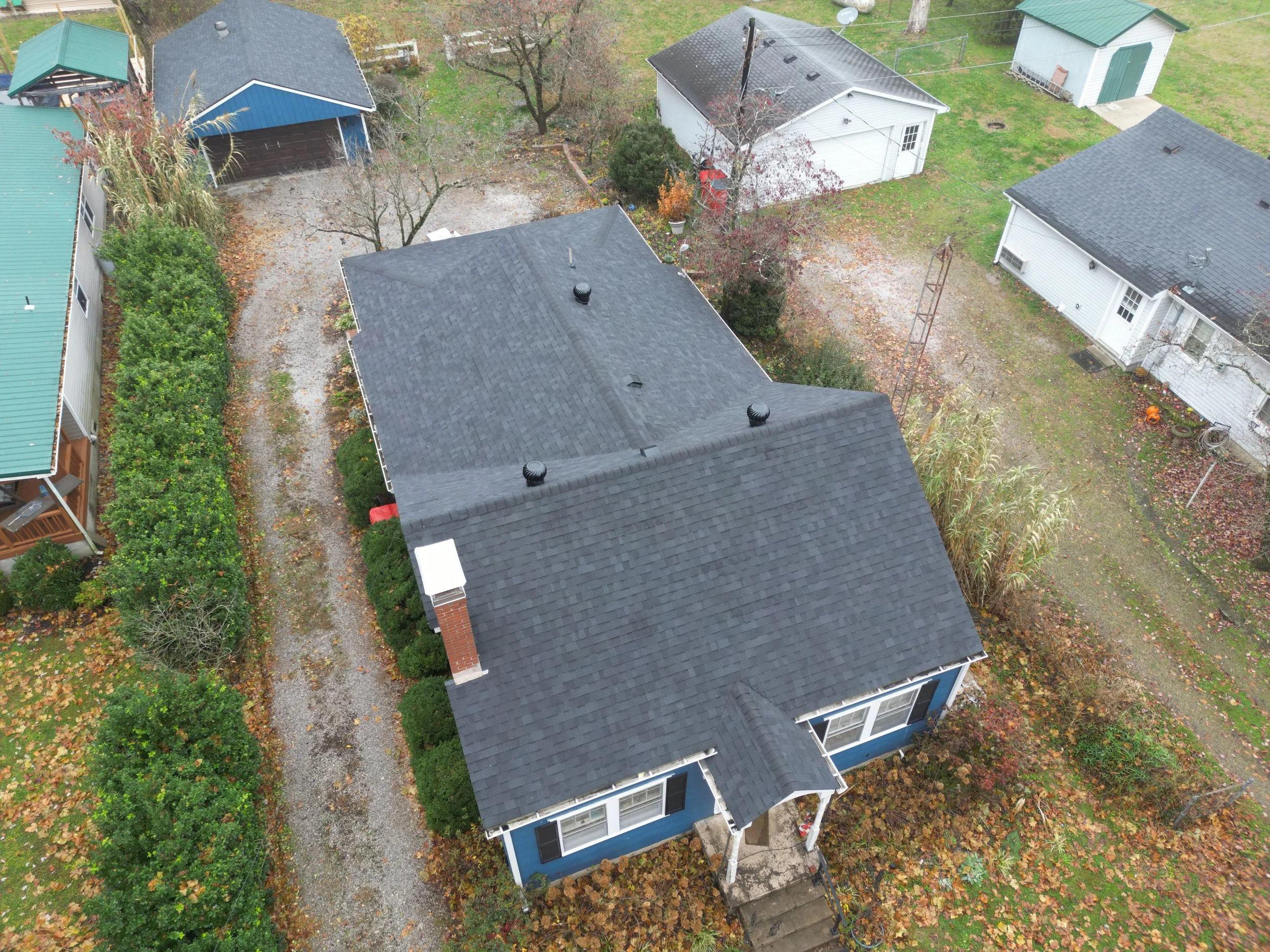 An aerial view of a house with a black roof, surrounded by other houses and trees on an autumn day with fallen leaves on the ground.