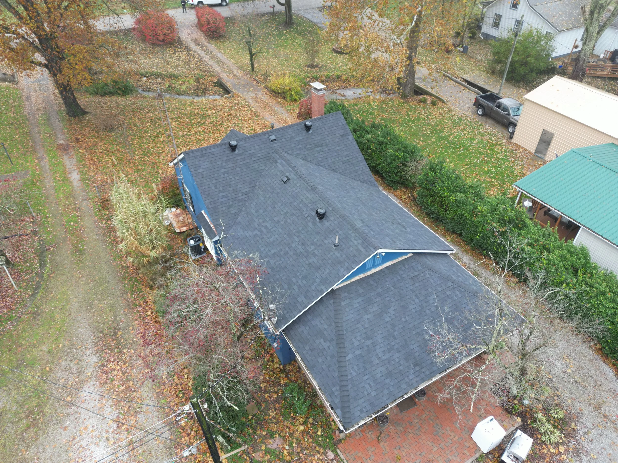 An aerial view of a house surrounded by trees with bare branches and a yard covered in fallen autumn leaves.