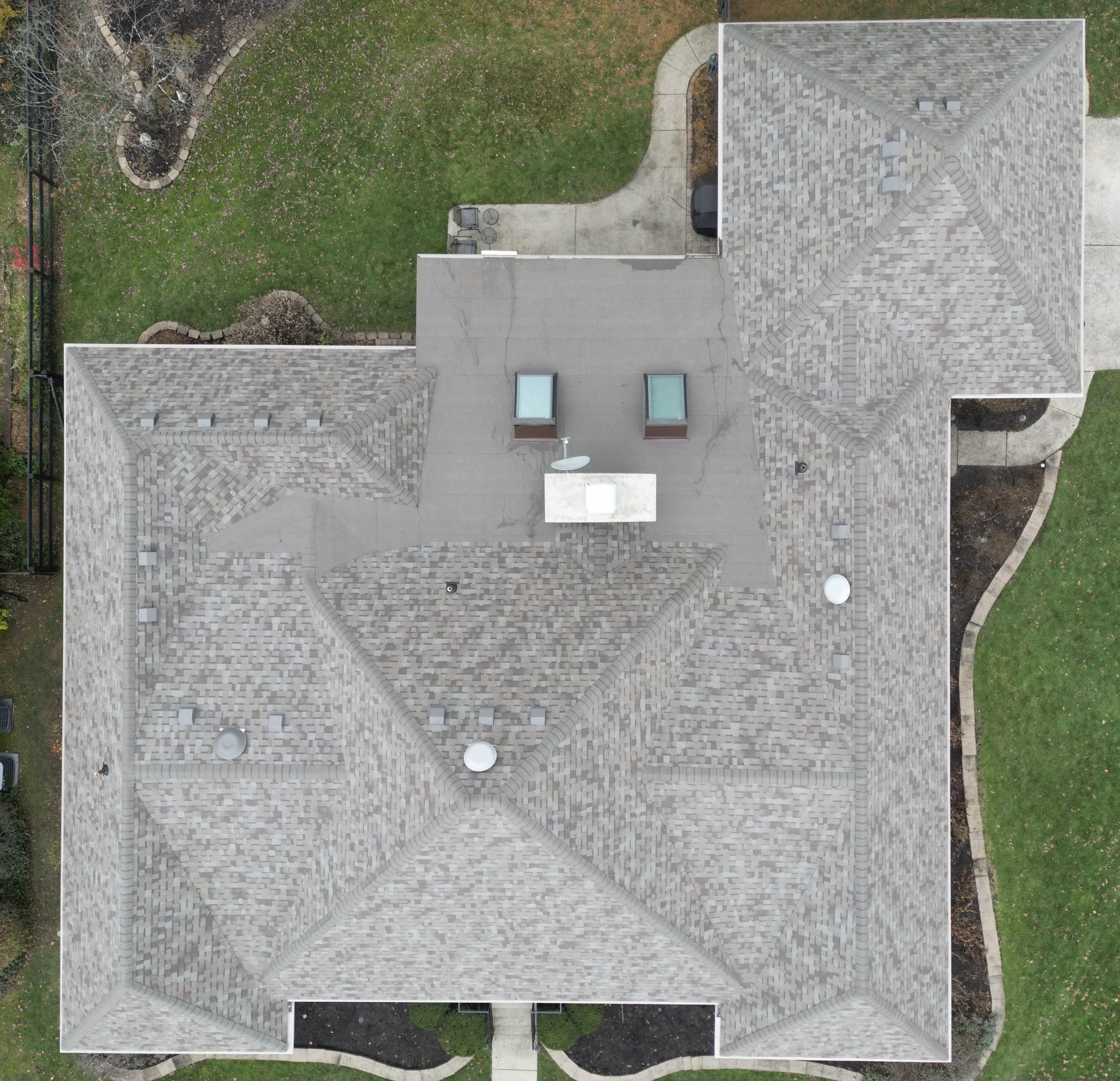 An aerial view of a house with a complex multi-section roof made of shingles, with various vents and skylights. Surrounding the house is a lawn with some plants and trees, a driveway with a parked vehicle, and landscaped areas with pathways and garde