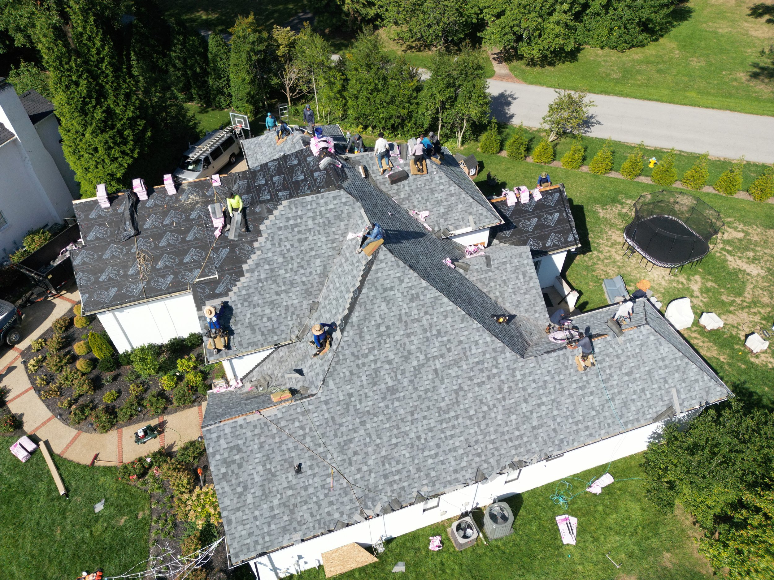 Aerial view of a house under construction with multiple workers installing roofing shingles on the roof. Tools, materials, and equipment are visible on the roof and surrounding yard.