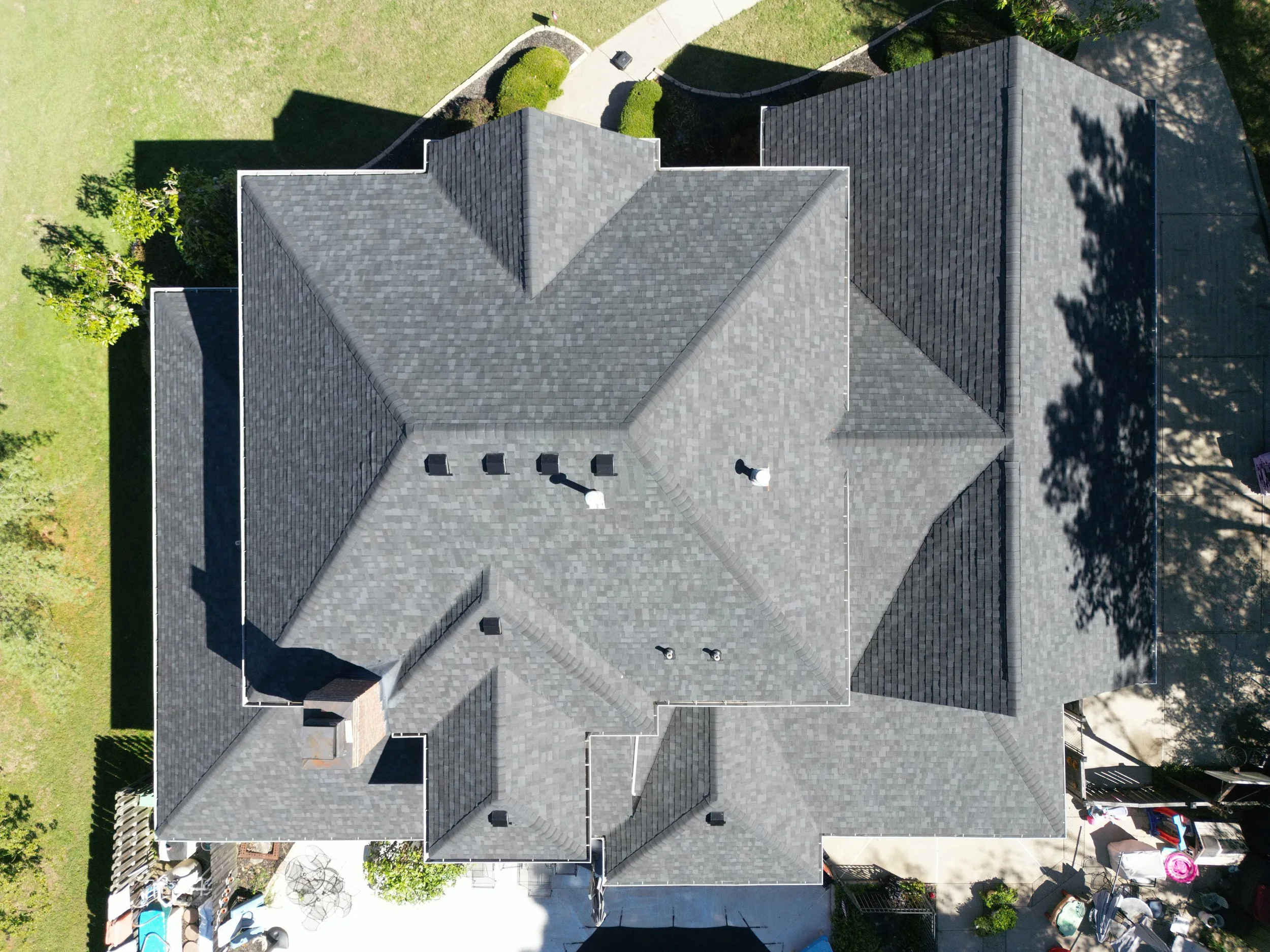 An aerial view of a gray shingled house roof with multiple peaks, vents, and chimneys, surrounded by green lawns and a driveway.
