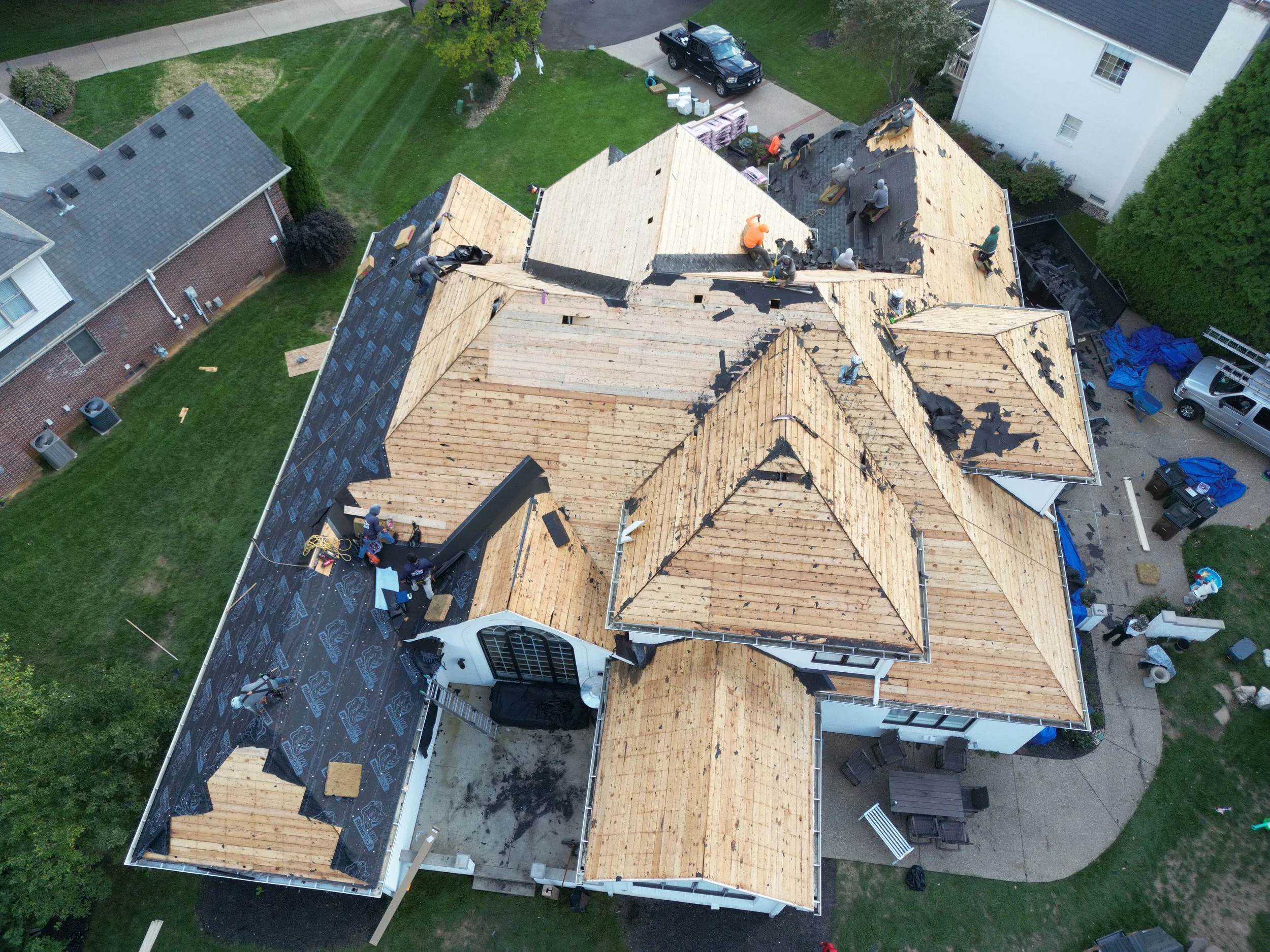 An aerial view of a large house under construction showing workers installing roofing with wooden panels and black roofing material. The house has multiple sections with different roof styles, and construction equipment and materials are visible arou