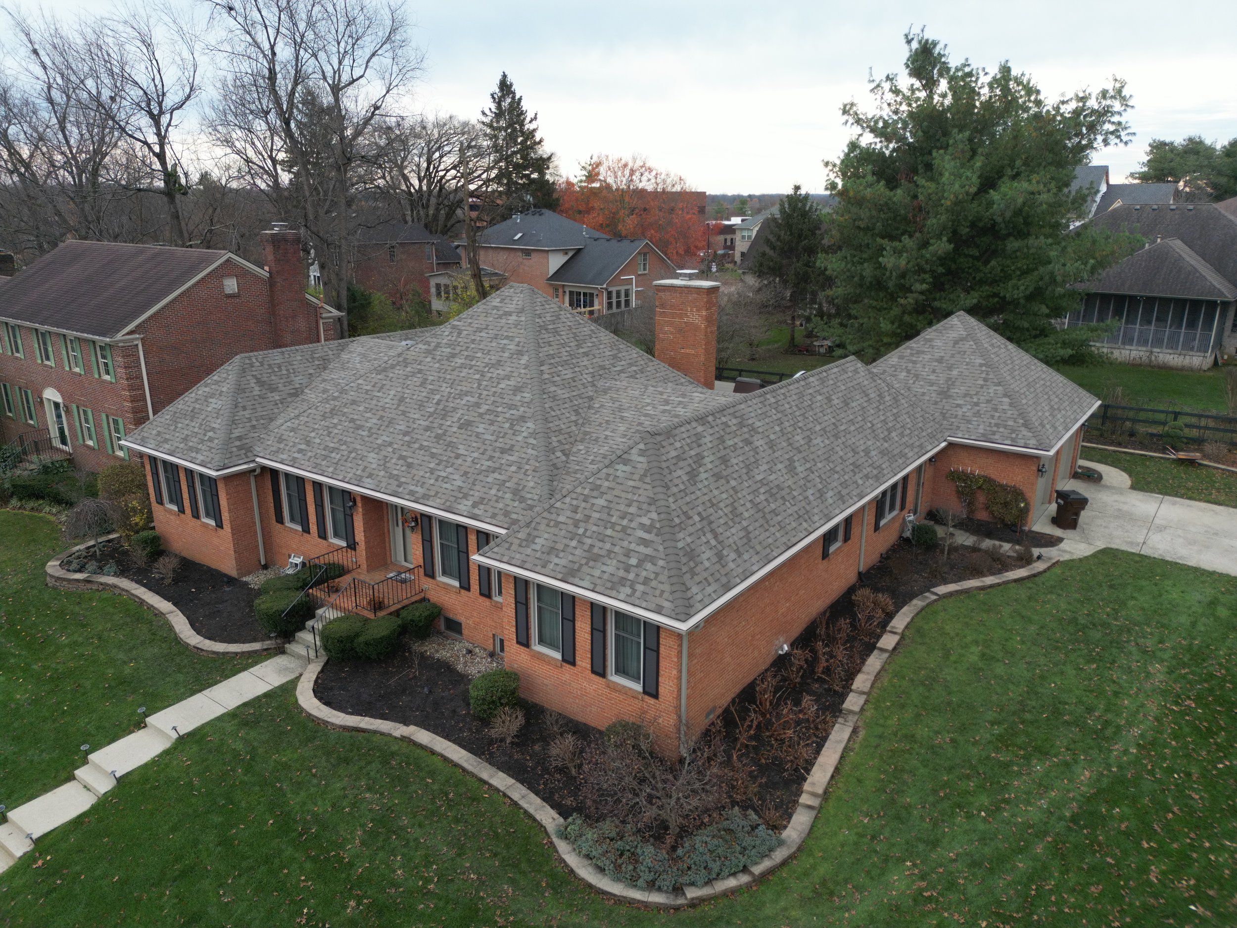 Aerial view of a brick house with a gray shingle roof, surrounded by a lawn, sidewalk, and other residential houses and trees in the background.
