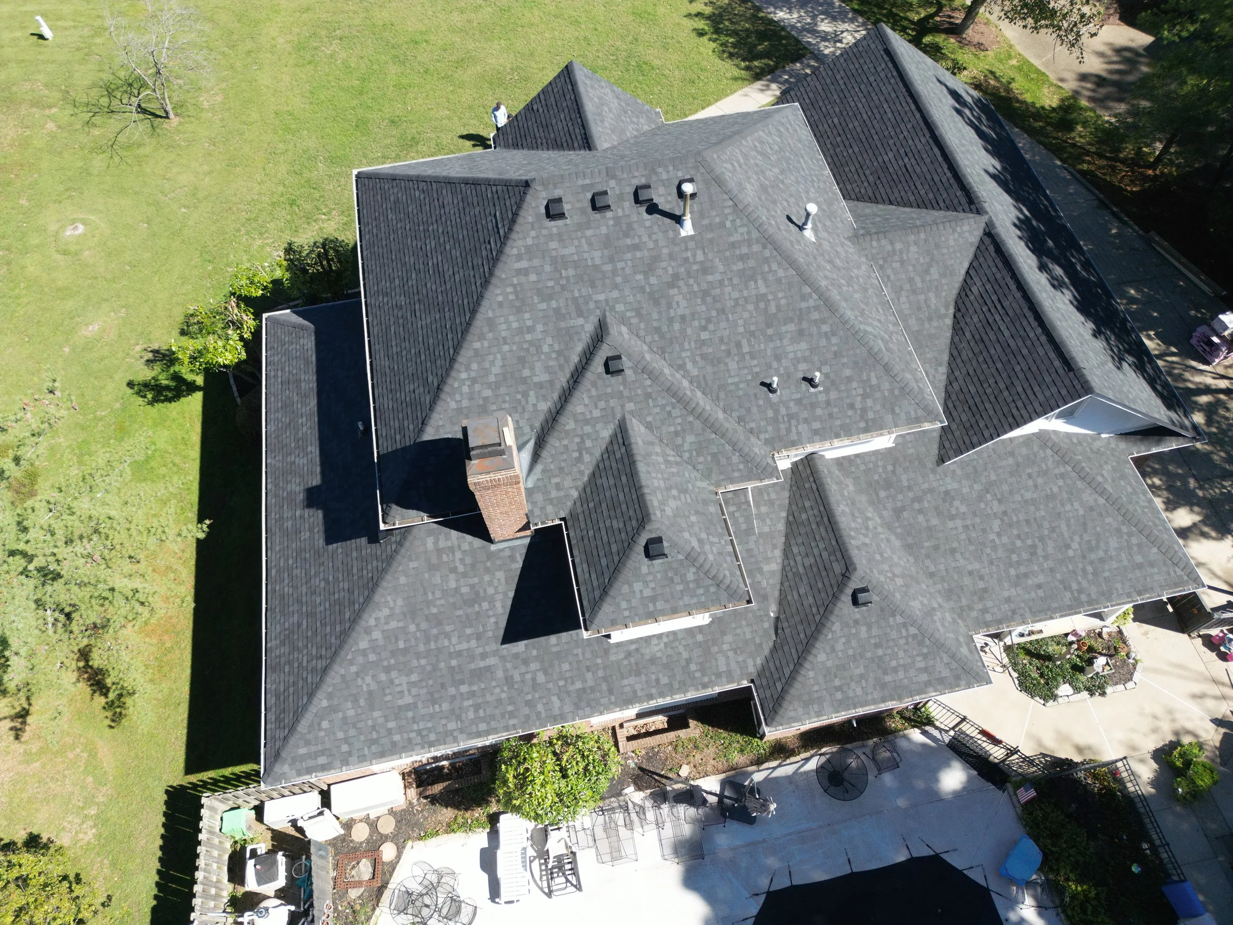 Aerial view of a house with a complex roof design covered in gray shingles, surrounded by a green yard with trees and a backyard patio area.