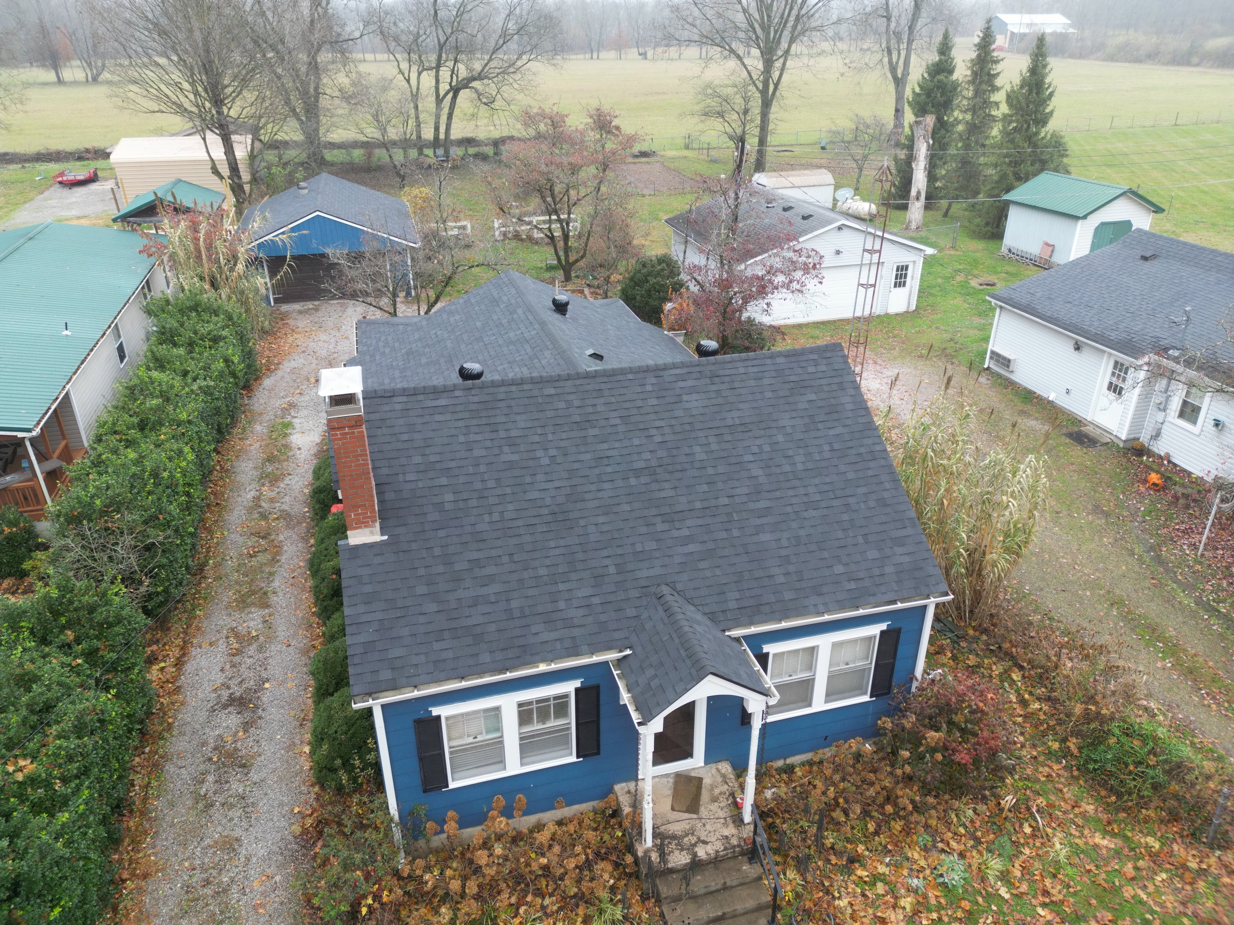 Aerial view of a blue house with a gray roof, surrounded by other houses, trees, and a grassy yard in a rural area.