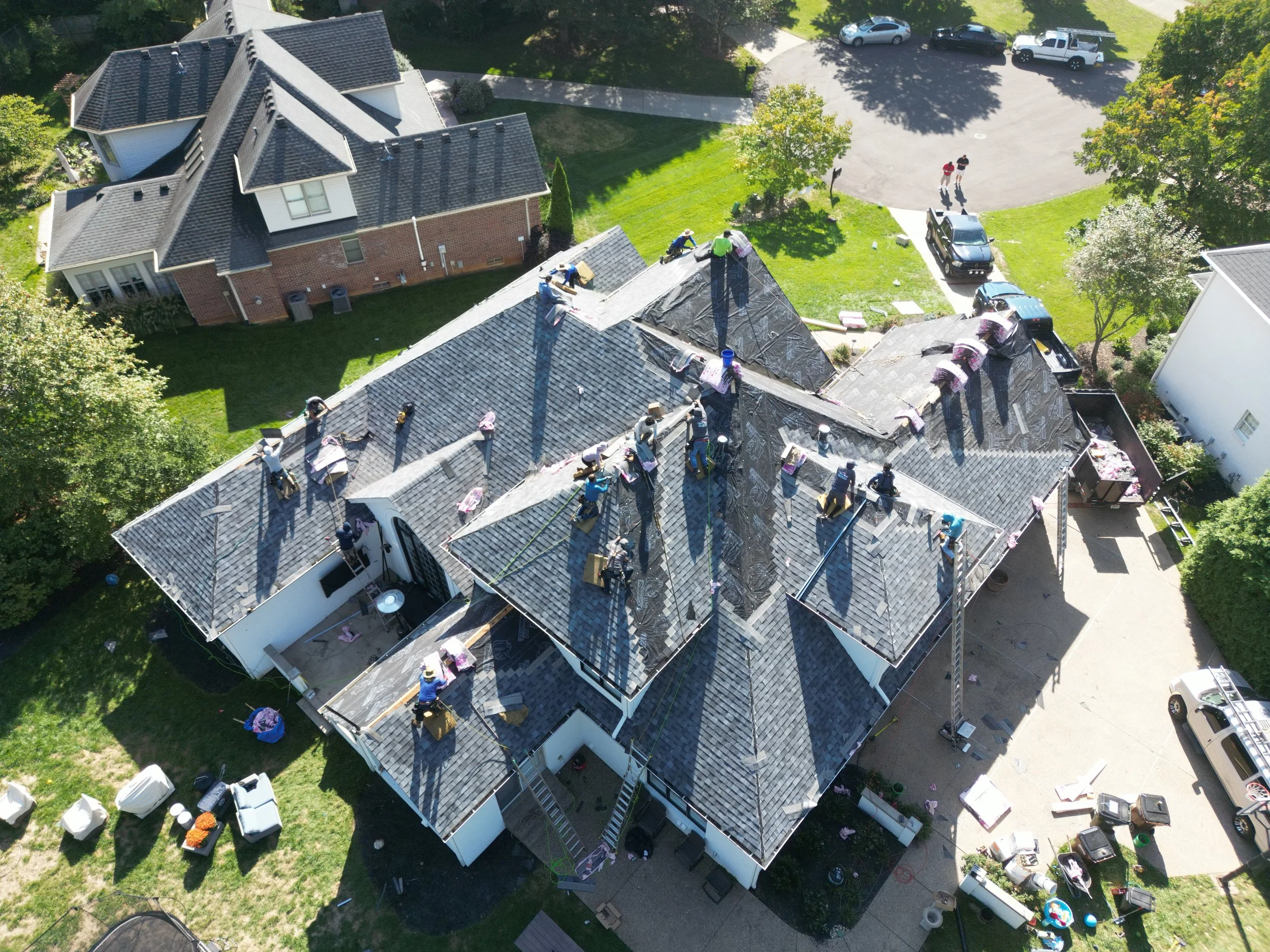 Aerial view of a house undergoing roof renovation, with workers on the roof installing new shingles, surrounded by green yard and cars parked in the driveway.