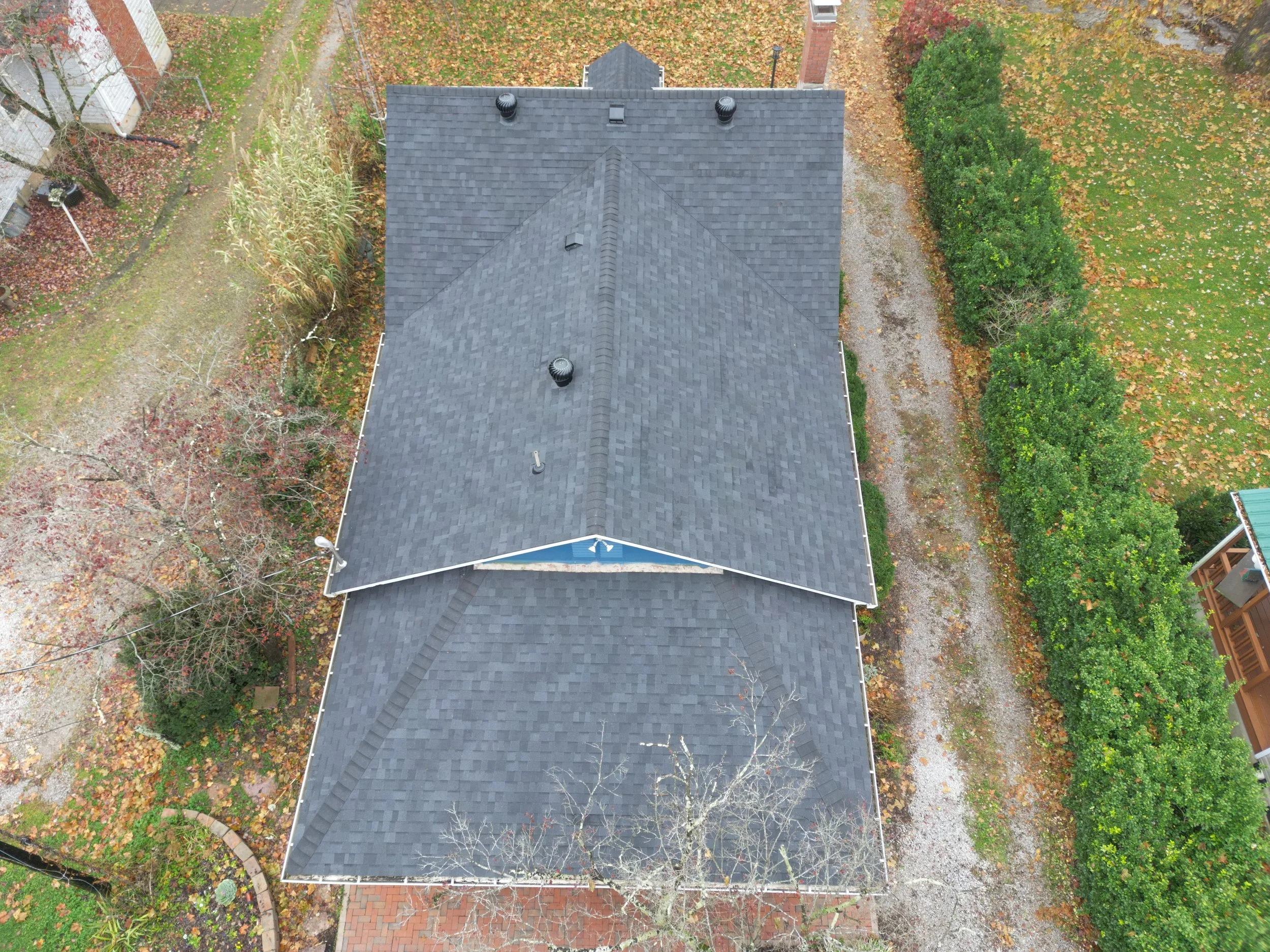 An aerial view of a house with a dark shingled roof, surrounded by trees, shrubs, and a yard with fallen autumn leaves.