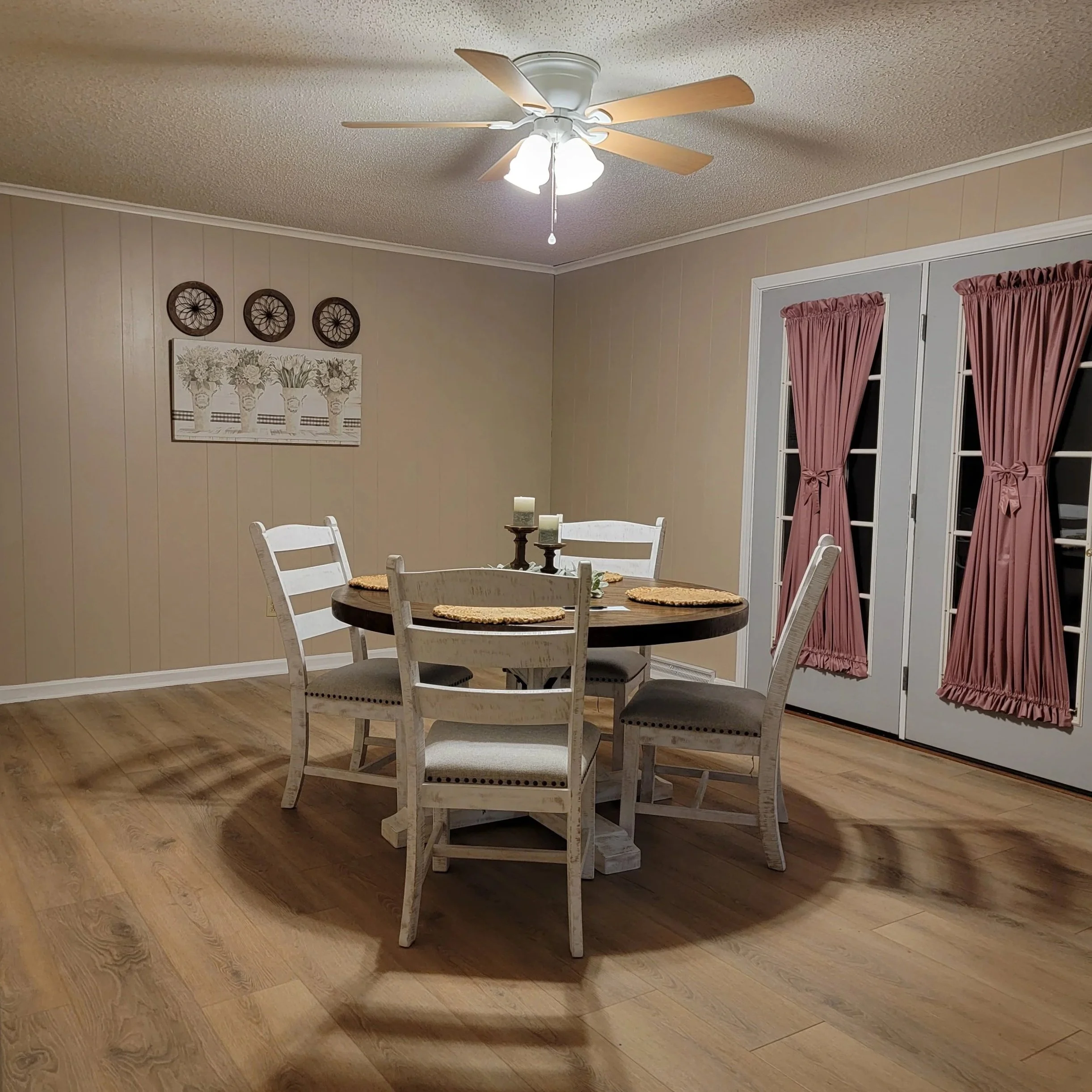 A dining room with a round wooden table and five white chairs with beige cushions. The table has three candles on stands and four woven placemats. There are pink curtains on glass doors and a wall decoration of three round dark-colored wall hangings above a flower-themed wall art. The room has beige paneled walls, a beige ceiling with a fan and light fixture, and wooden flooring.