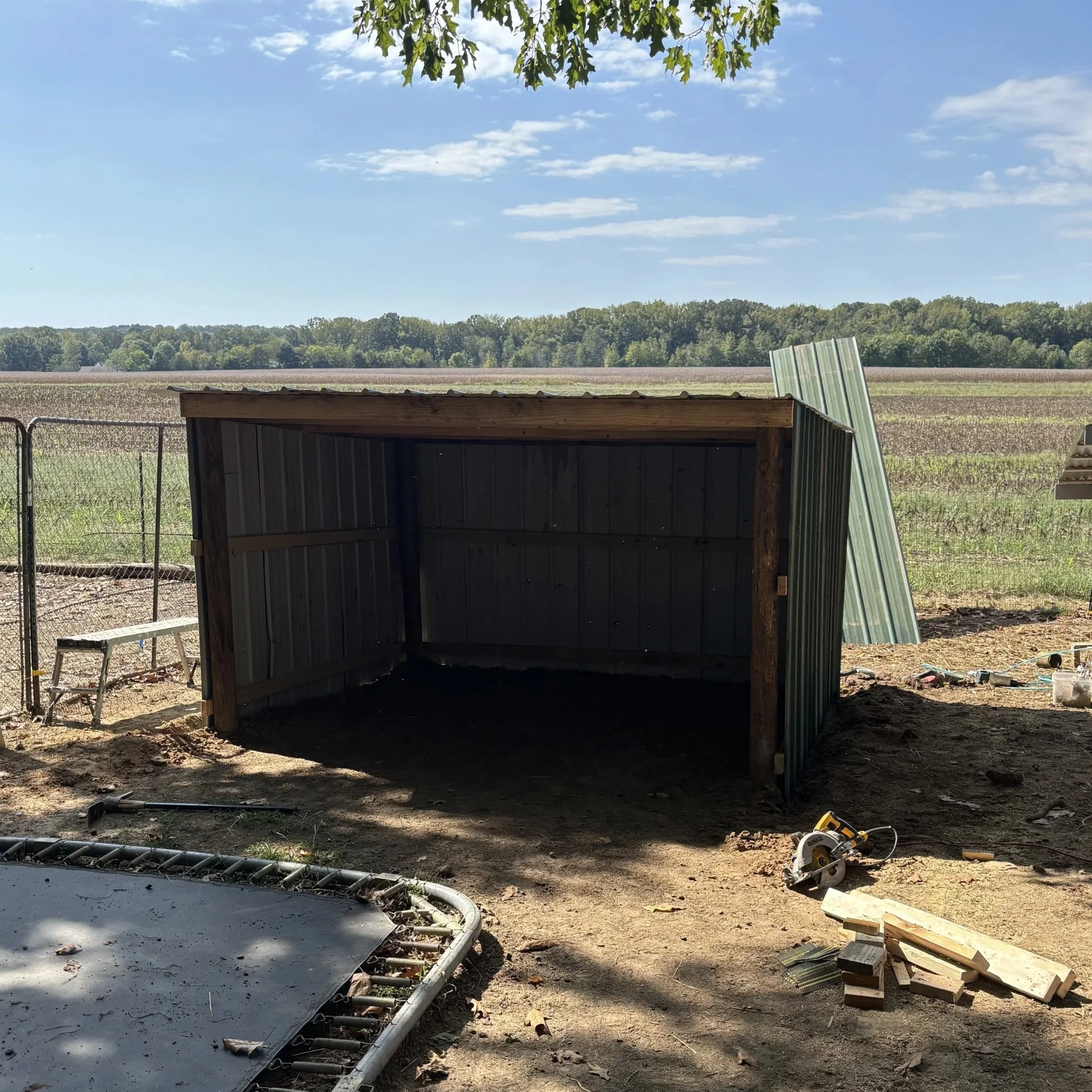 A partially constructed shed with metal walls in an outdoor setting, with tools and construction materials scattered around, and a large open field in the background under a partly cloudy sky.
