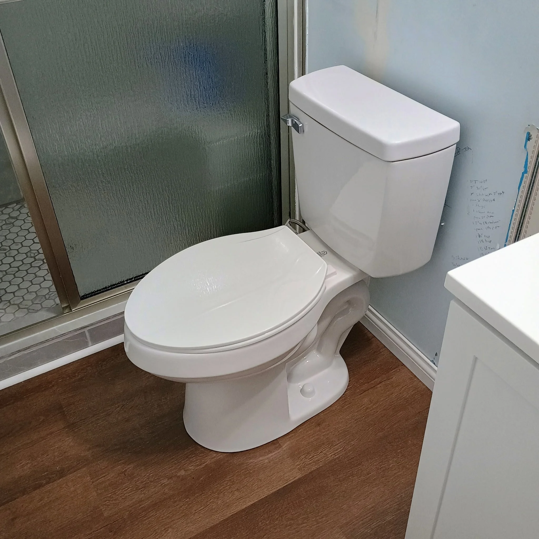 A white toilet next to a glass shower door with textured glass and a metal frame, on a wooden floor in a bathroom.