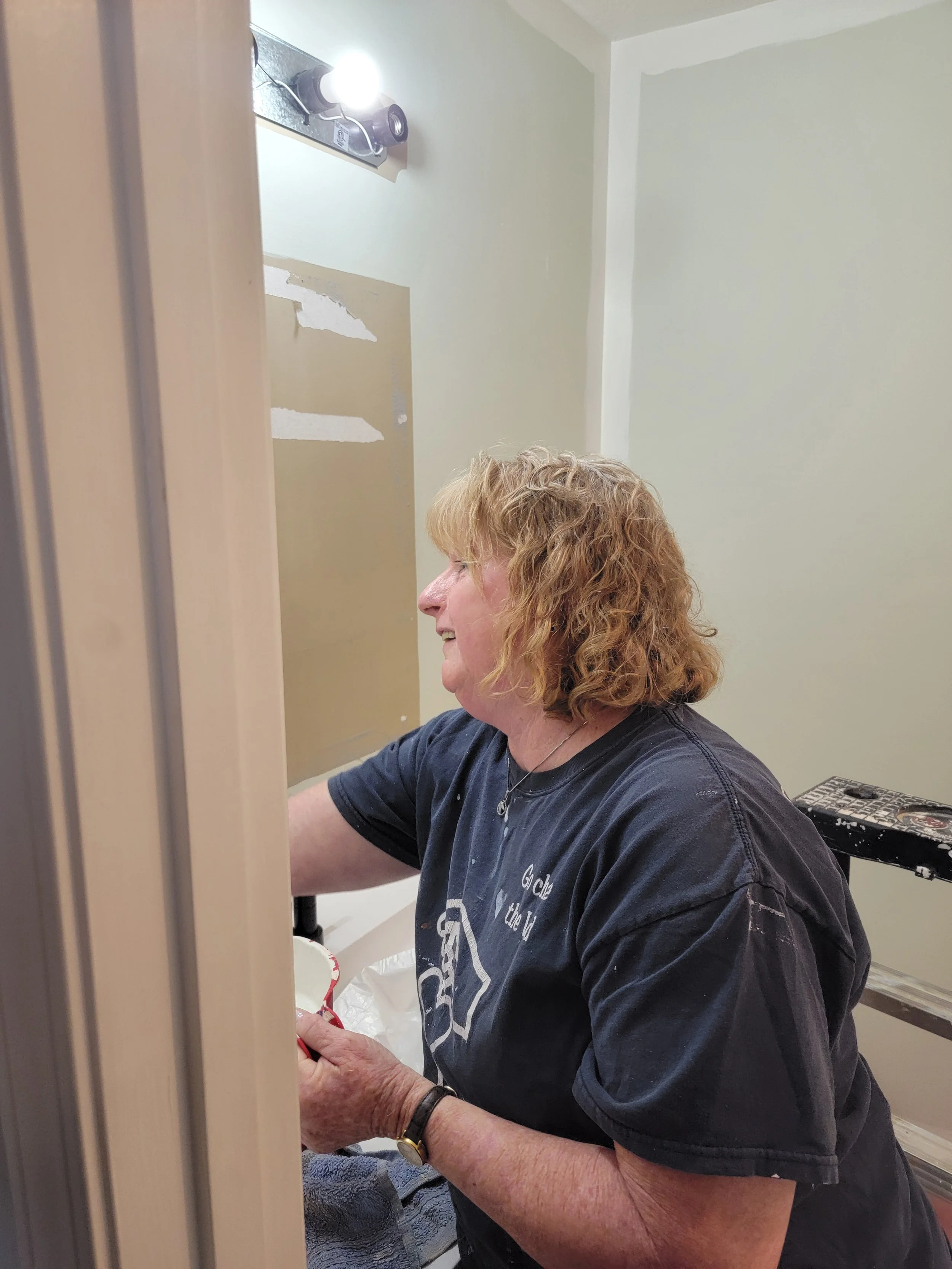 A woman with curly blond hair and a black T-shirt working on a wall near a mirror with a bright light above her, on a stepladder.