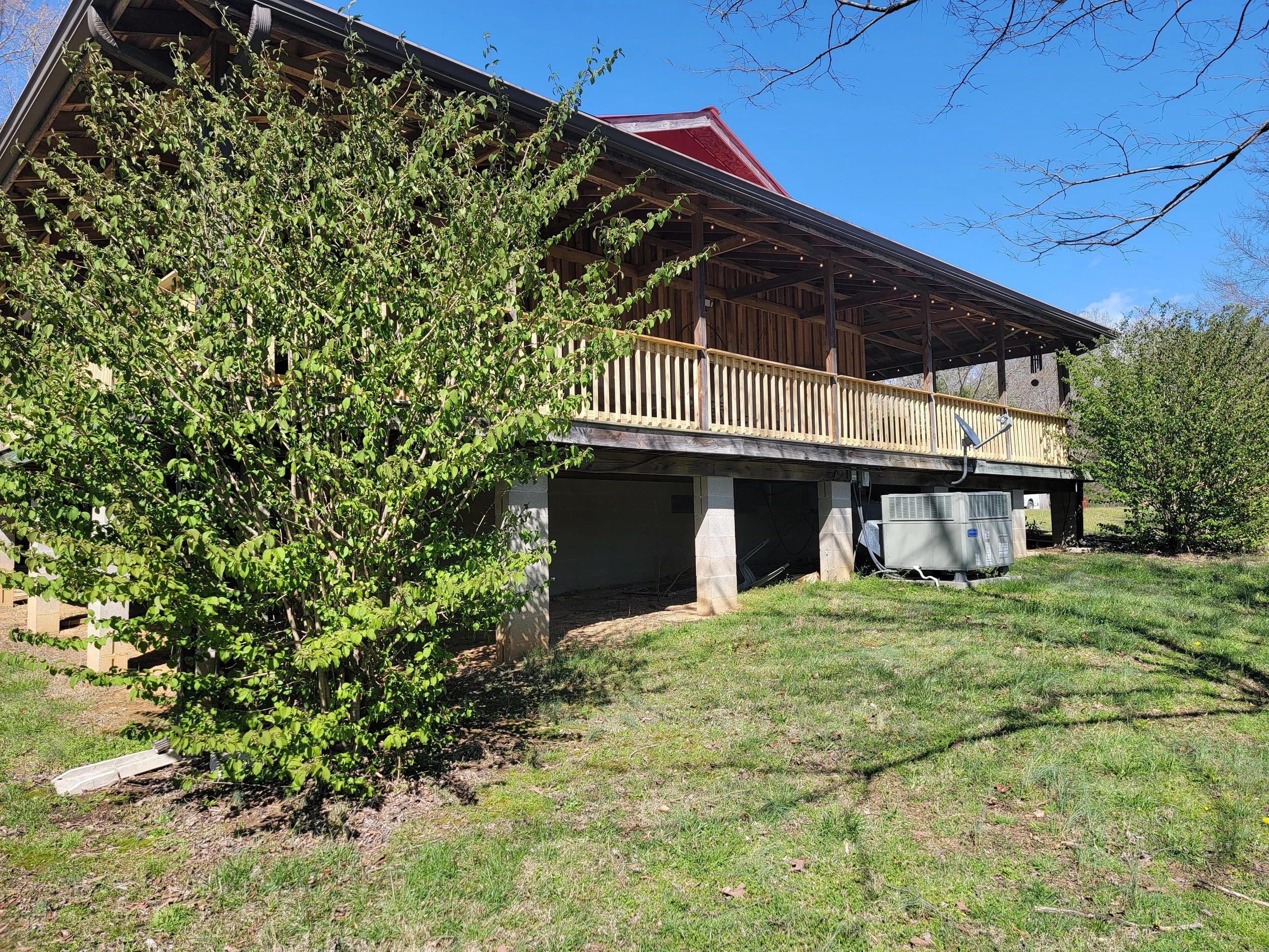 A backyard view of a house with a wooden deck and railing, raised on stilts. There are two green bushes, one on the left and one on the right, with a grassy area in the foreground. A clear blue sky and leafless tree branches are visible in the backgr
