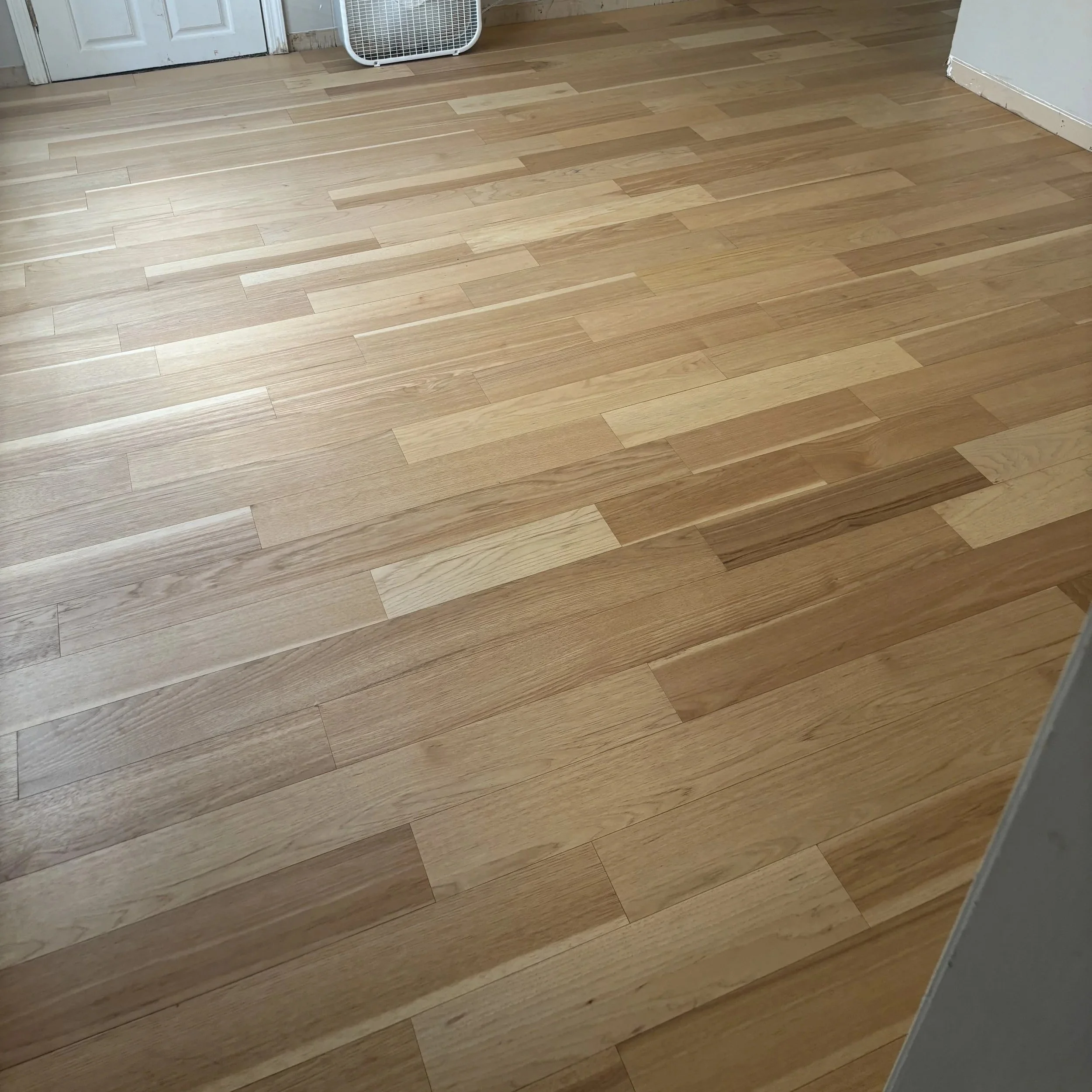 A hardwood floor with light natural wood finish, showing the grain and variation in planks, in a room with white walls and a partially visible white door and fan.